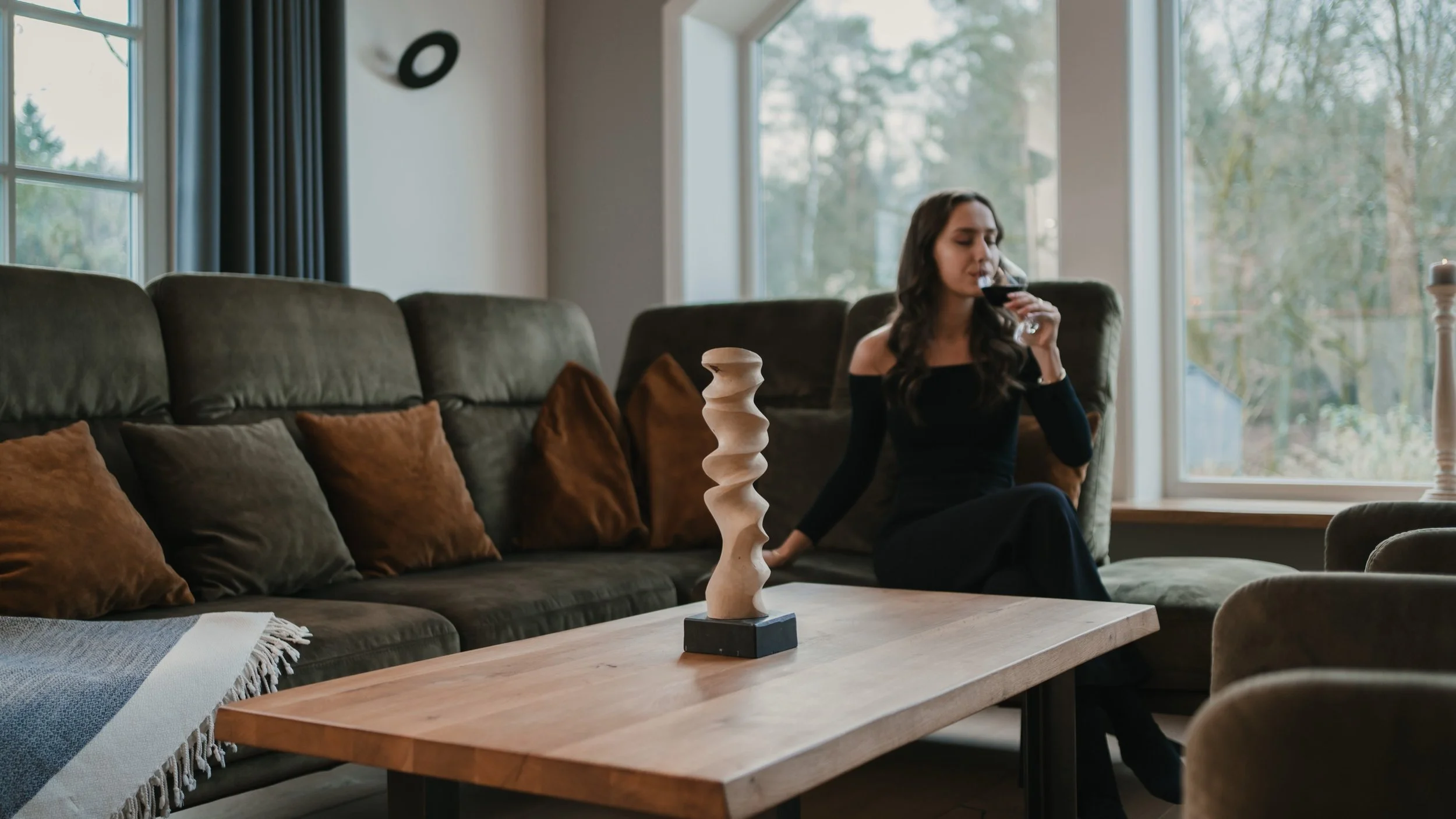 Eine Frau sitzt auf einem dunkelgrünen Sofa, trinkt Rotwein und schaut nach draußen, in einer modern eingerichteten Wohnzimmer mit großen Fenstern. Auf dem Tisch steht eine skulpturartige Holzkreation.