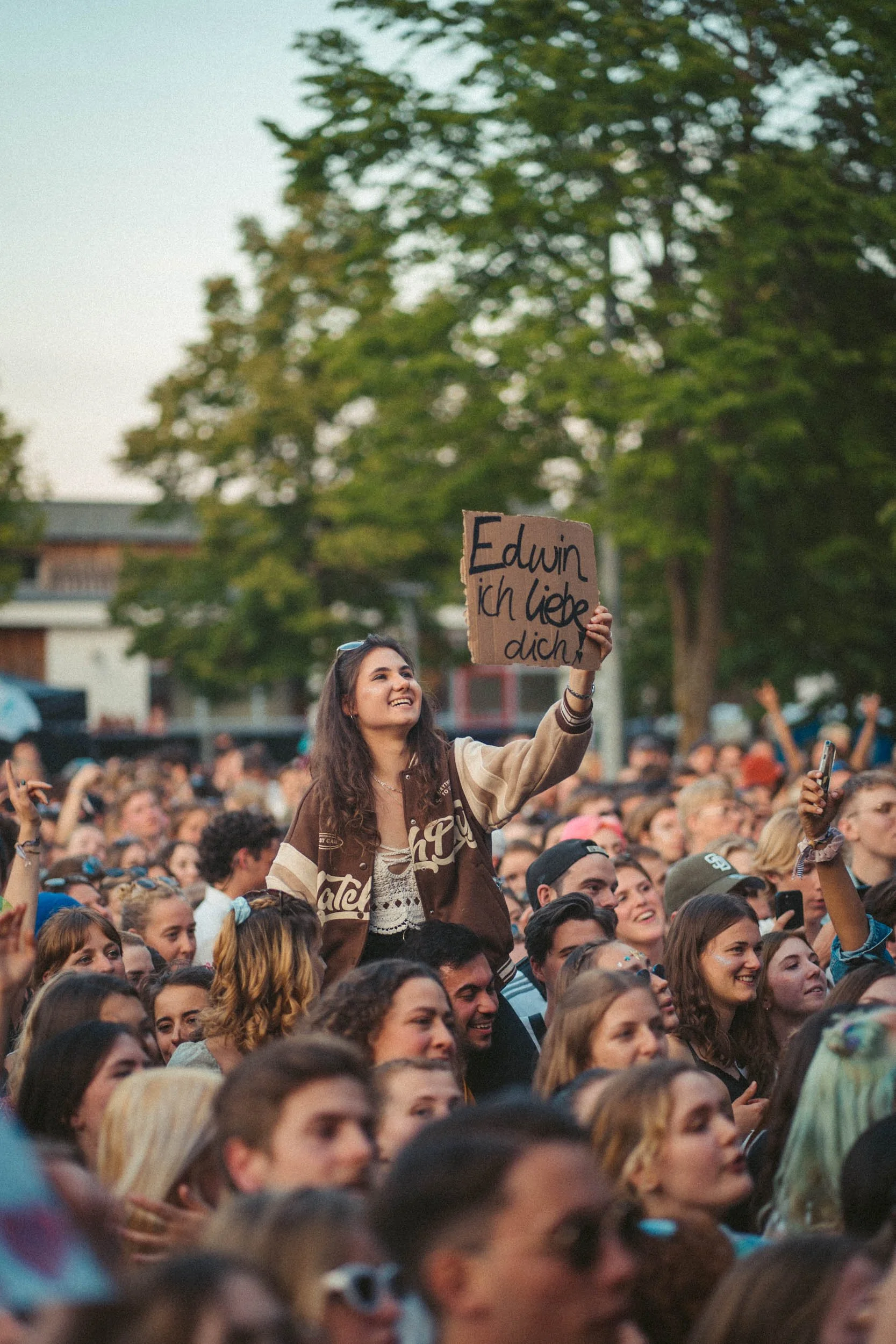 Eine junge Frau hält ein Schild mit der Aufschrift 'Edwin ich liebe dich' inmitten einer Menschenmenge bei einem Konzert oder Festival im Freien.