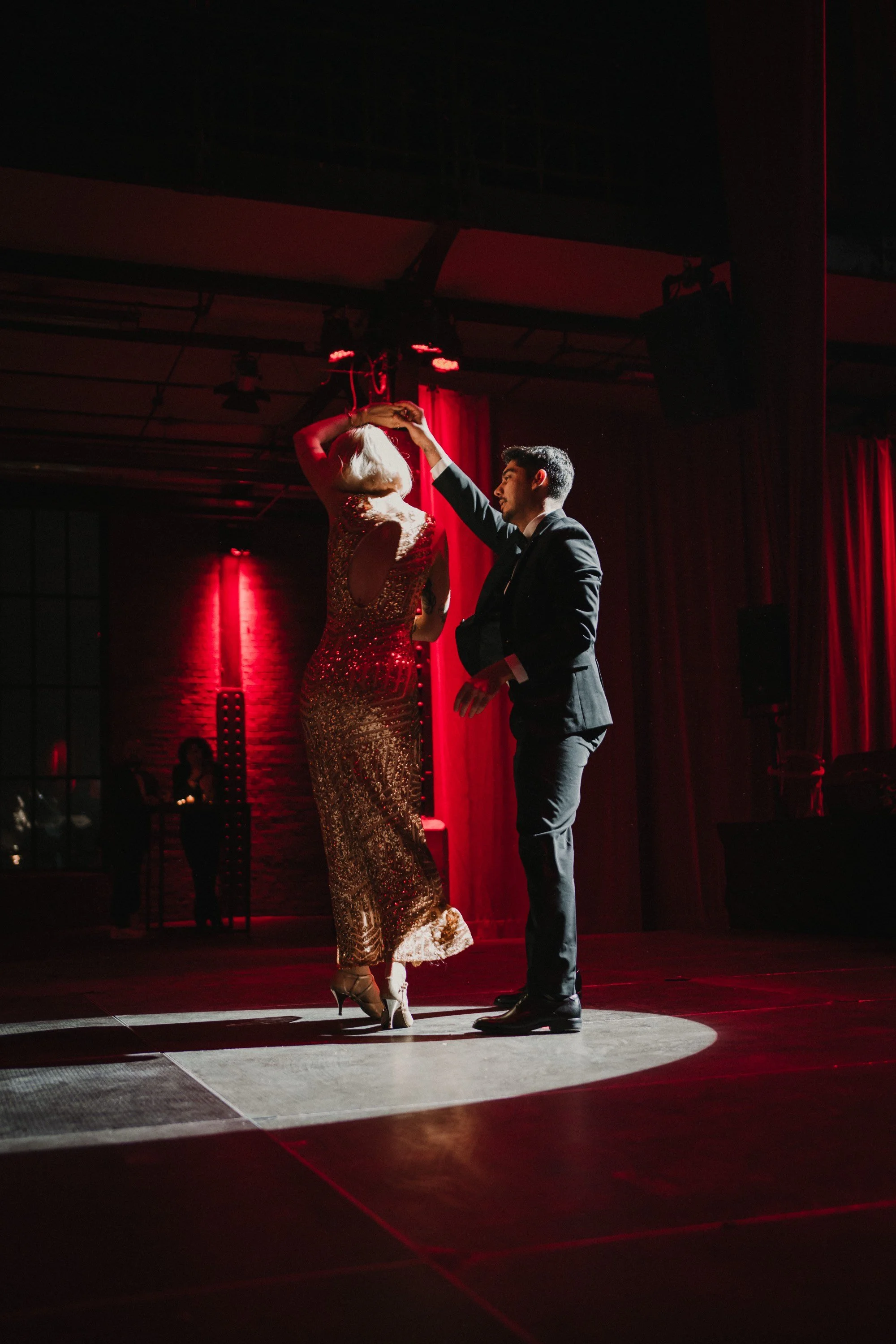 Ein Mann und eine Frau tanzen auf einer Bühne, mit rotem Hintergrund und Scheinwerferlicht, in eleganter Kleidung, beim Tanz im Rahmen einer Veranstaltung.