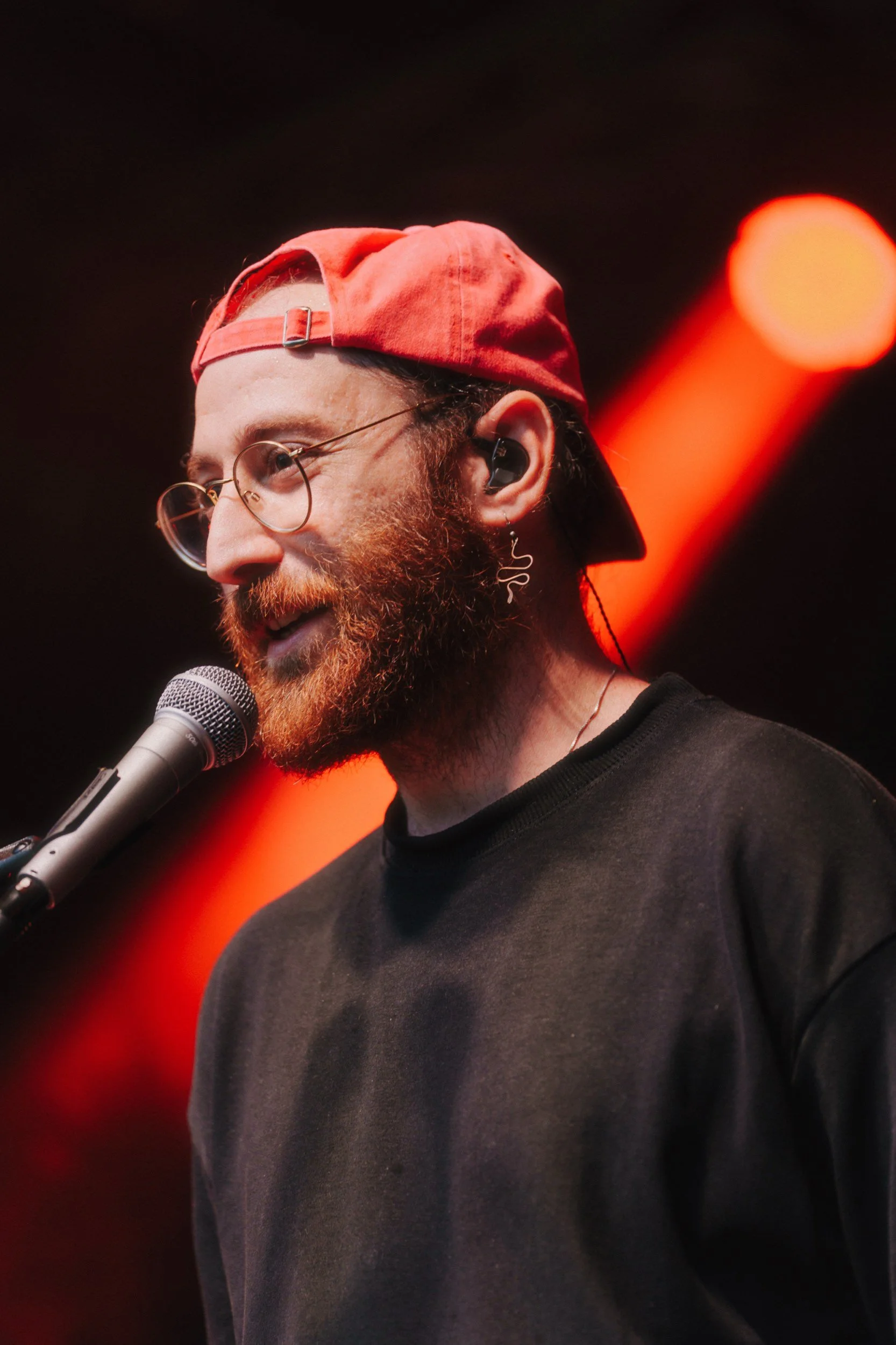 Ein Mann mit rotbonem Baseballcap, Brille, Barthaare und Earpiece spricht in ein Mikrofon bei einem Konzert oder einer Veranstaltung, im Hintergrund sind rote Lichter.