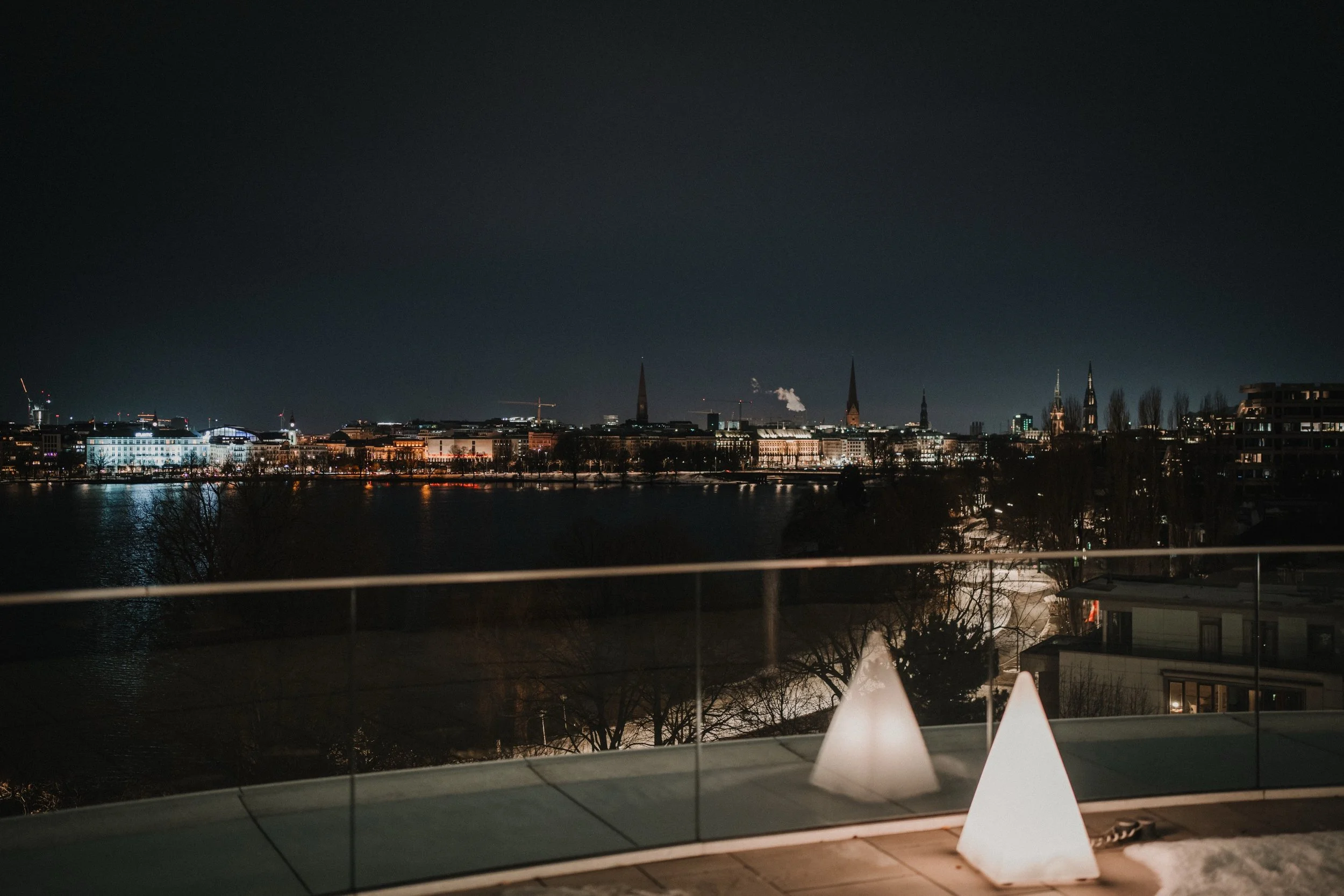 A nächtliche Stadtansicht mit beleuchteten Gebäuden über einem Fluss, im Vordergrund eine Terrasse mit beleuchteten Dekoelementen.
