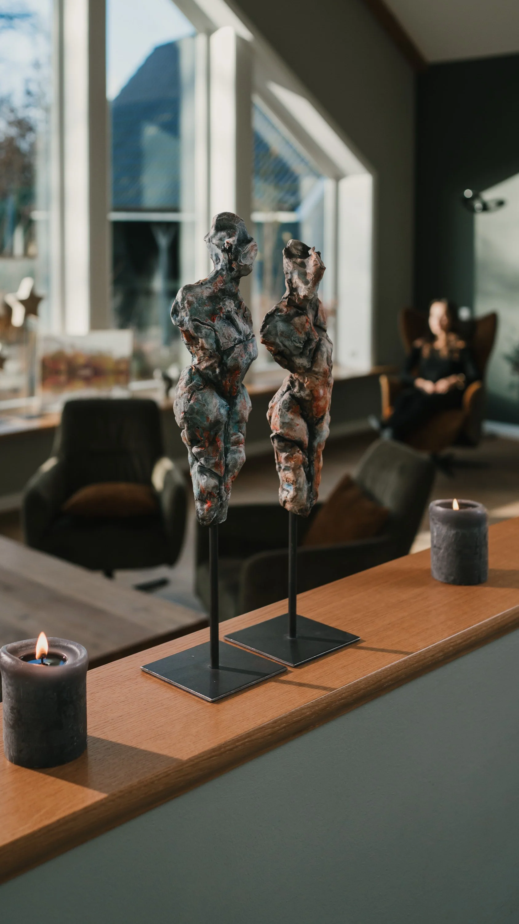 Zwei künstlerische Skulpturen, abstrakt gestaltete weibliche Figuren auf schwarzen Ständern, auf einem Holzregal, mit Kerzen davor.