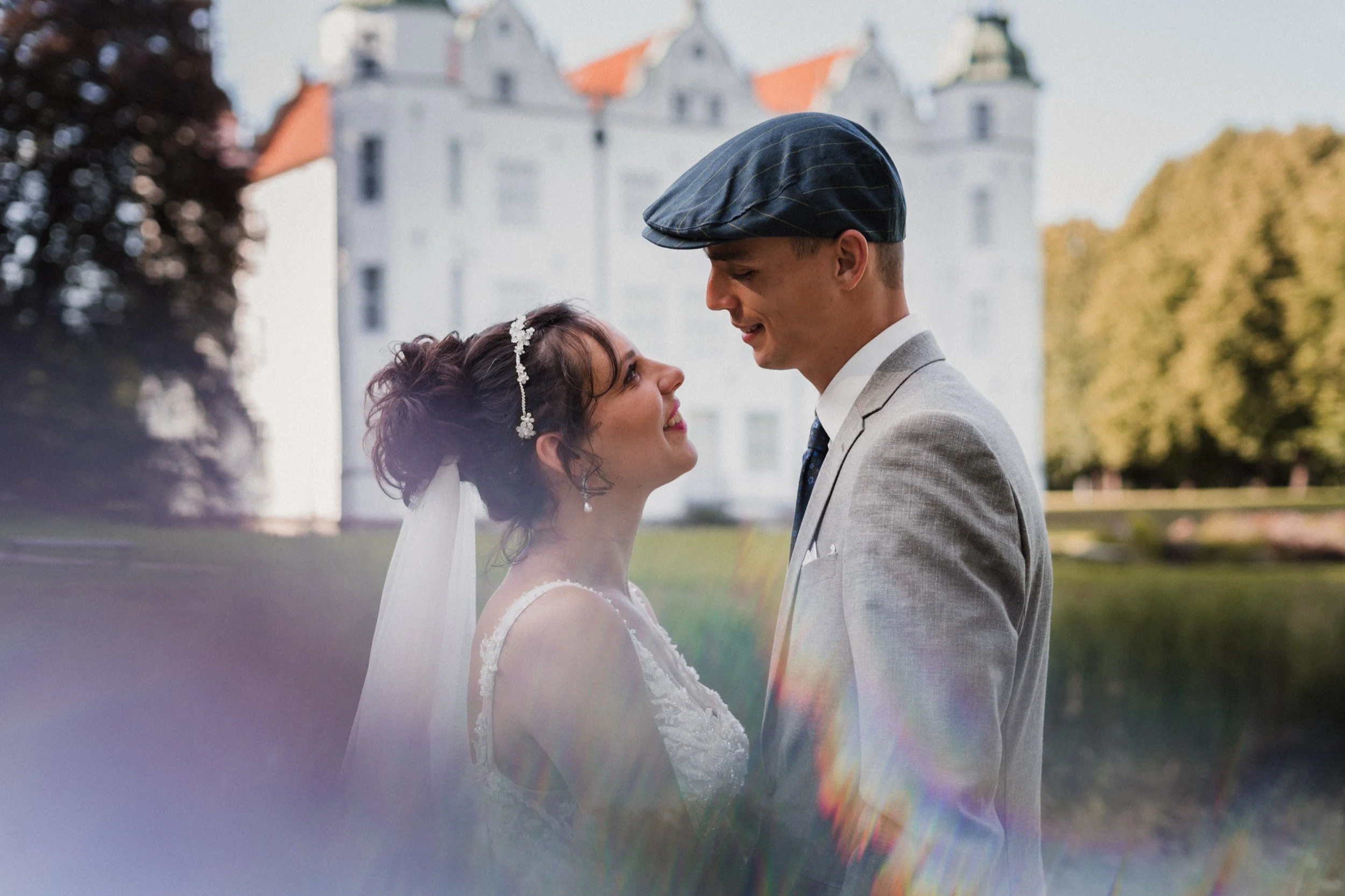 Ein Brautpaar auf einer Hochzeit steht sich gegenüber vor einem Schloss im Freien, umgeben von Bäumen und Rasen, während sie liebevoll in die Augen schauen.