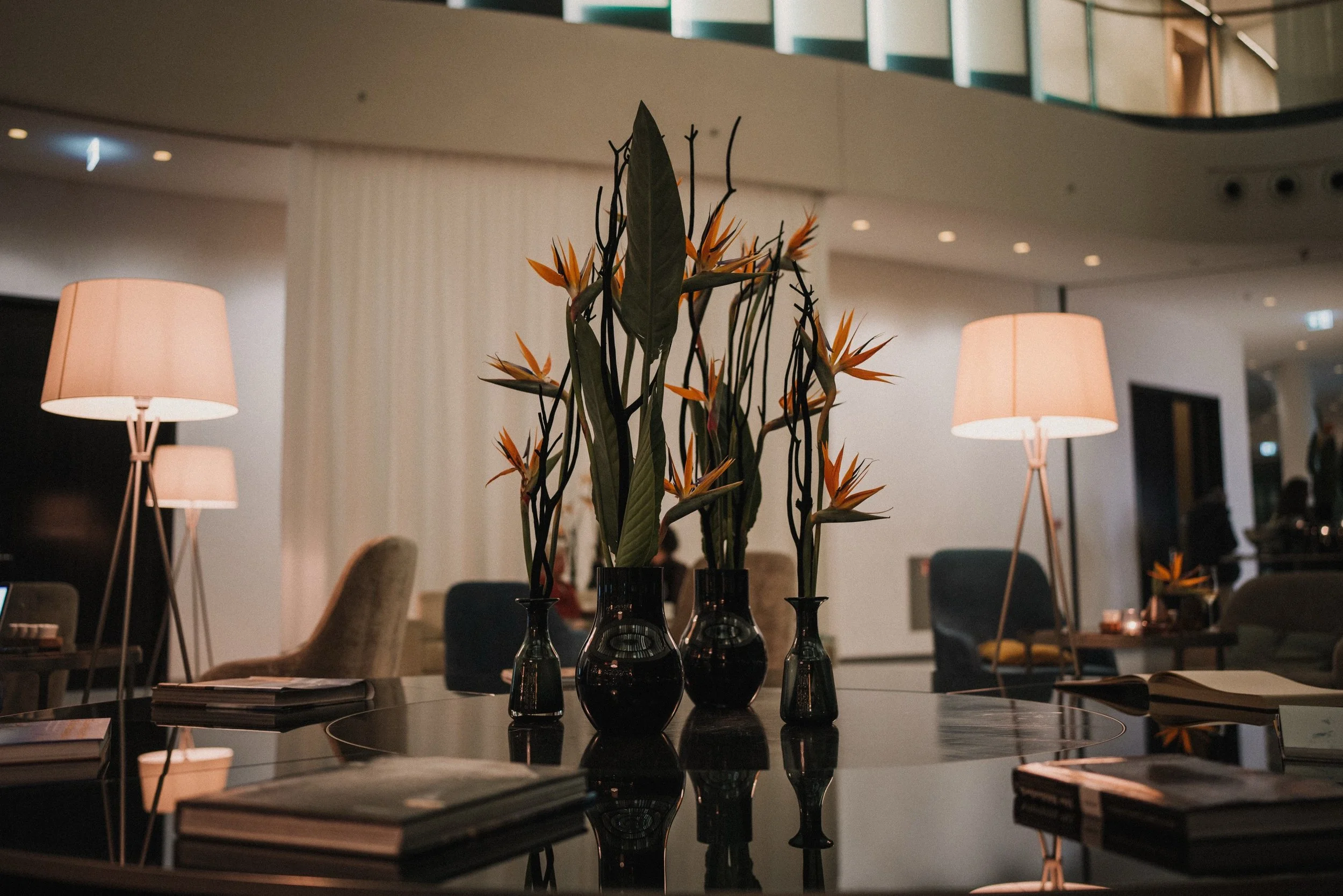 Ein stilvoller Innenraum mit einem Tisch, auf dem drei schwarze Vasen mit orangefarbenen Blumen stehen, umgeben von Stehlampen und bequemen Stühlen.