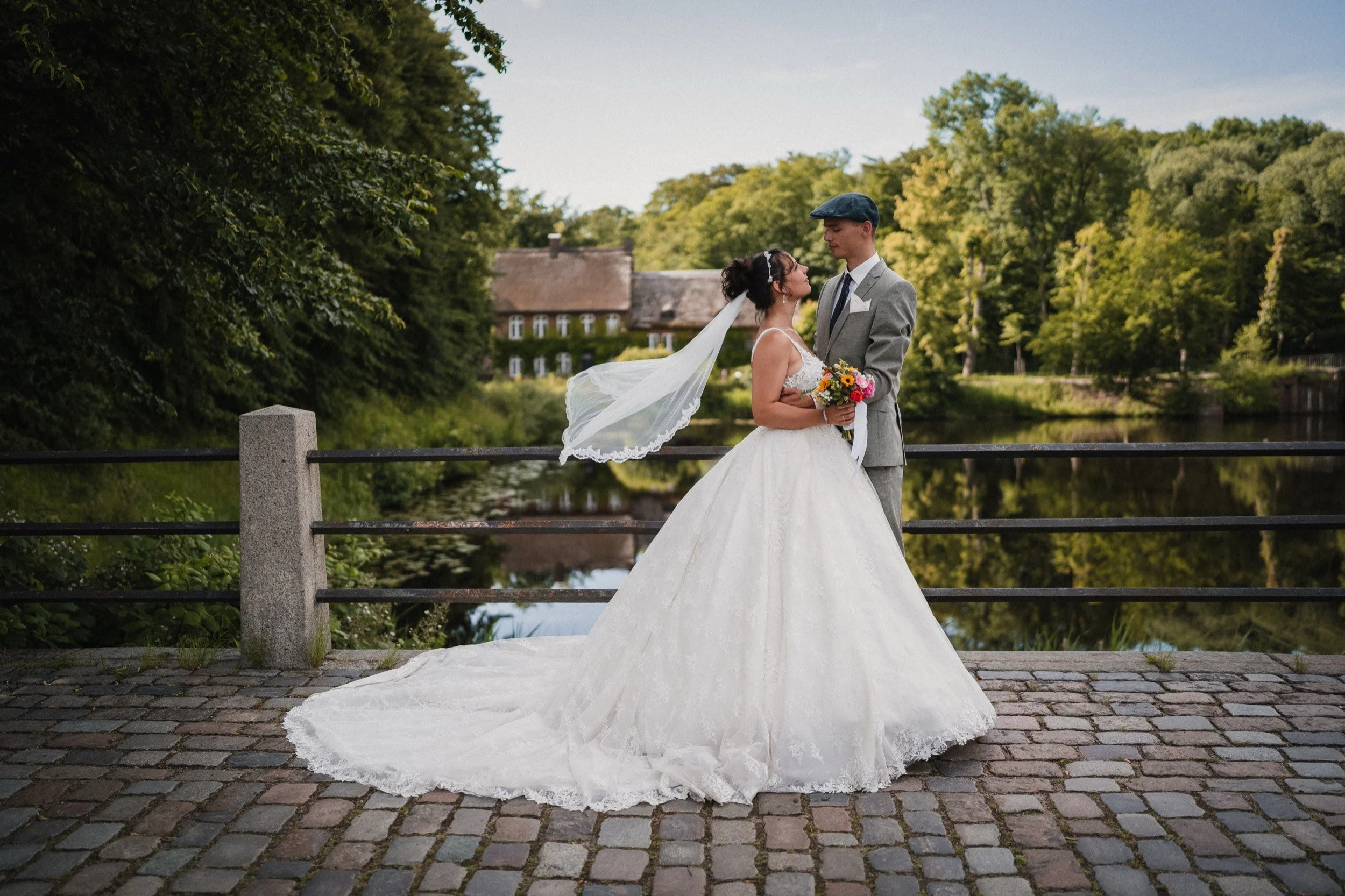 Ein Brautpaar steht sich auf einer Brücke gegenüber, umgeben von Bäumen, beim Sonnenuntergang, mit einem Fluss im Hintergrund.