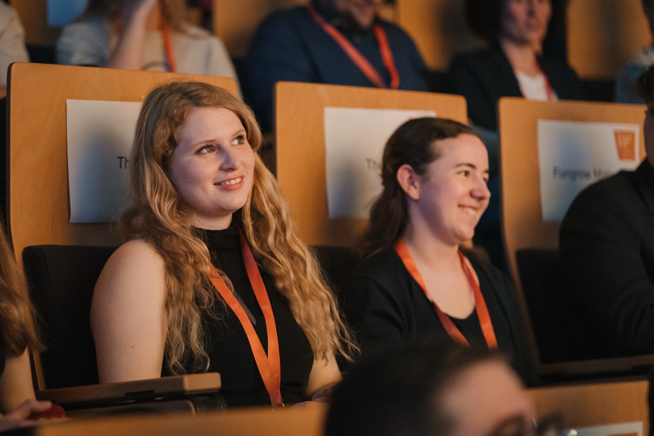 Zwei Frauen sitzen in einem dunklen Auditorium, lachen und schauen nach vorne, mit Lanyards um den Hals.