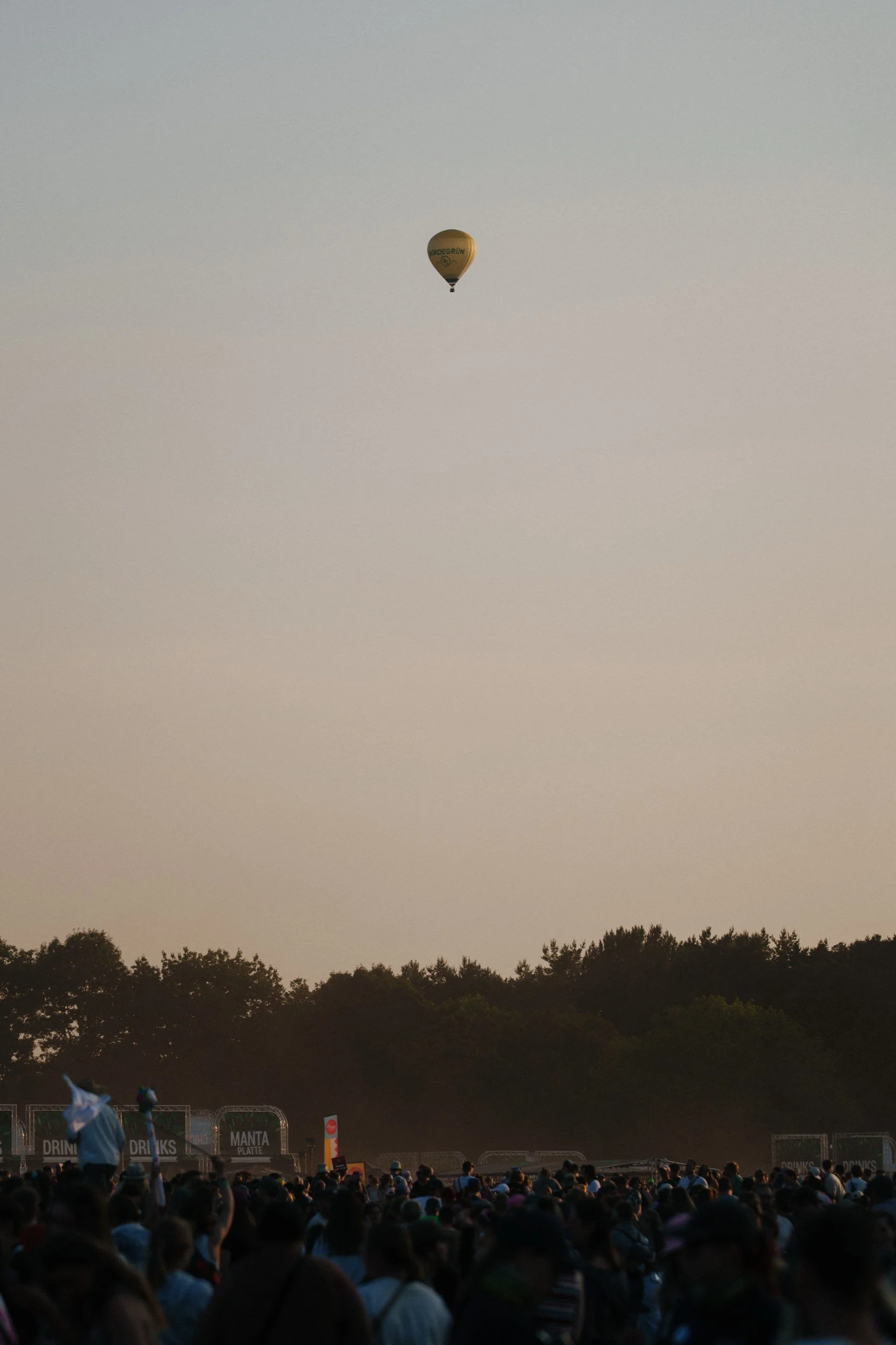 Ein Heißluftballon fliegt über einem Menschenmenge bei Dämmerlicht, umgeben von Bäumen.