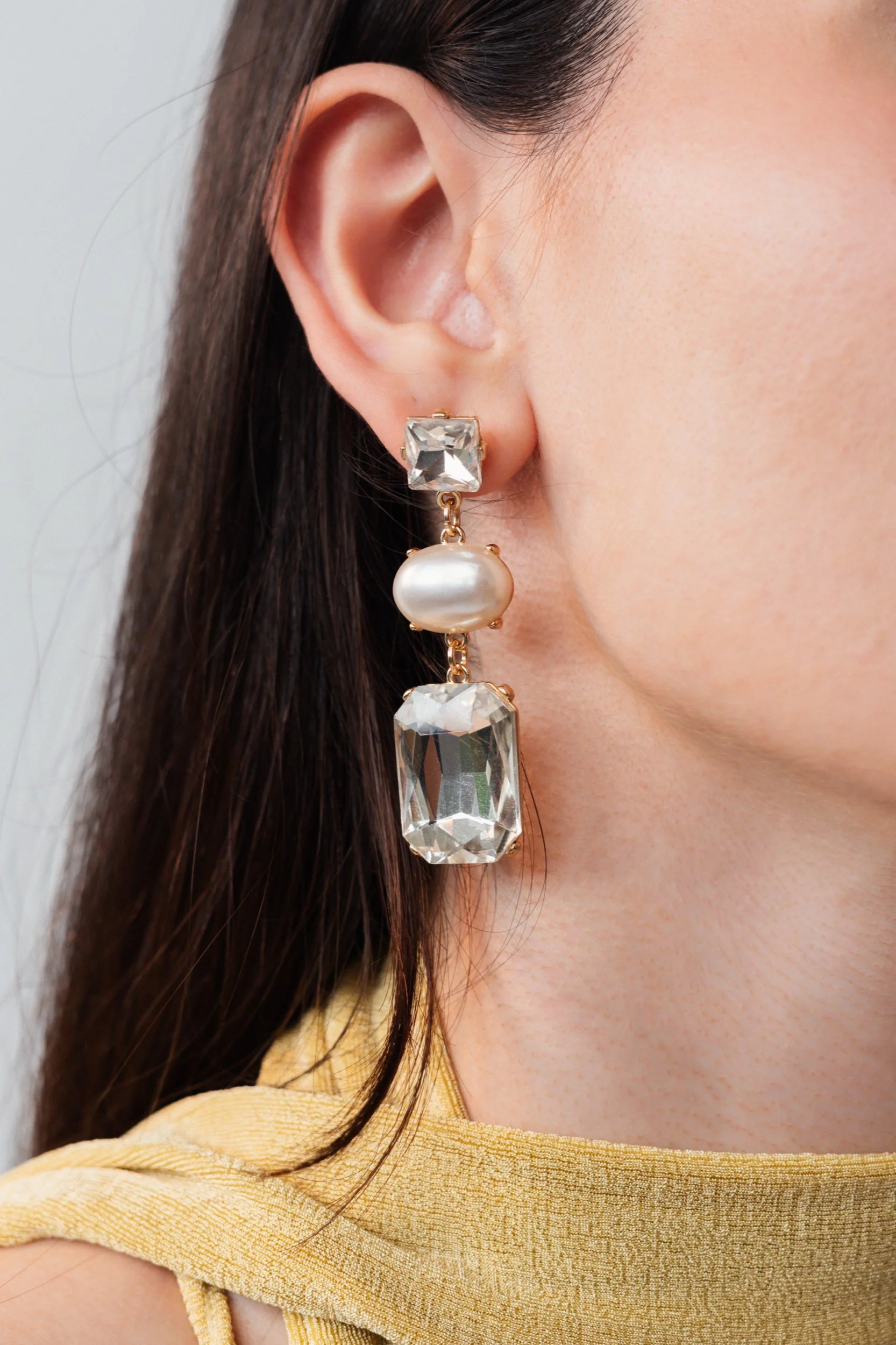 Nahaufnahme eines Ohrschmucks, bestehend aus einem großen Kristall, einer Perle und einem weiteren Kristall, getragen von einer Frau mit dunklen Haaren und gelbem Kleid.