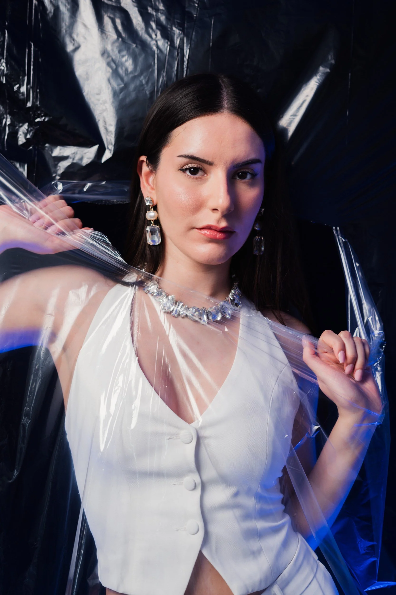 Porträt einer Frau, die schwarze Haare, Schmuck und eine weiße Jacke trägt, vor einem schwarzen und blauen Hintergrund, umgeben von durchsichtigem Plastik.