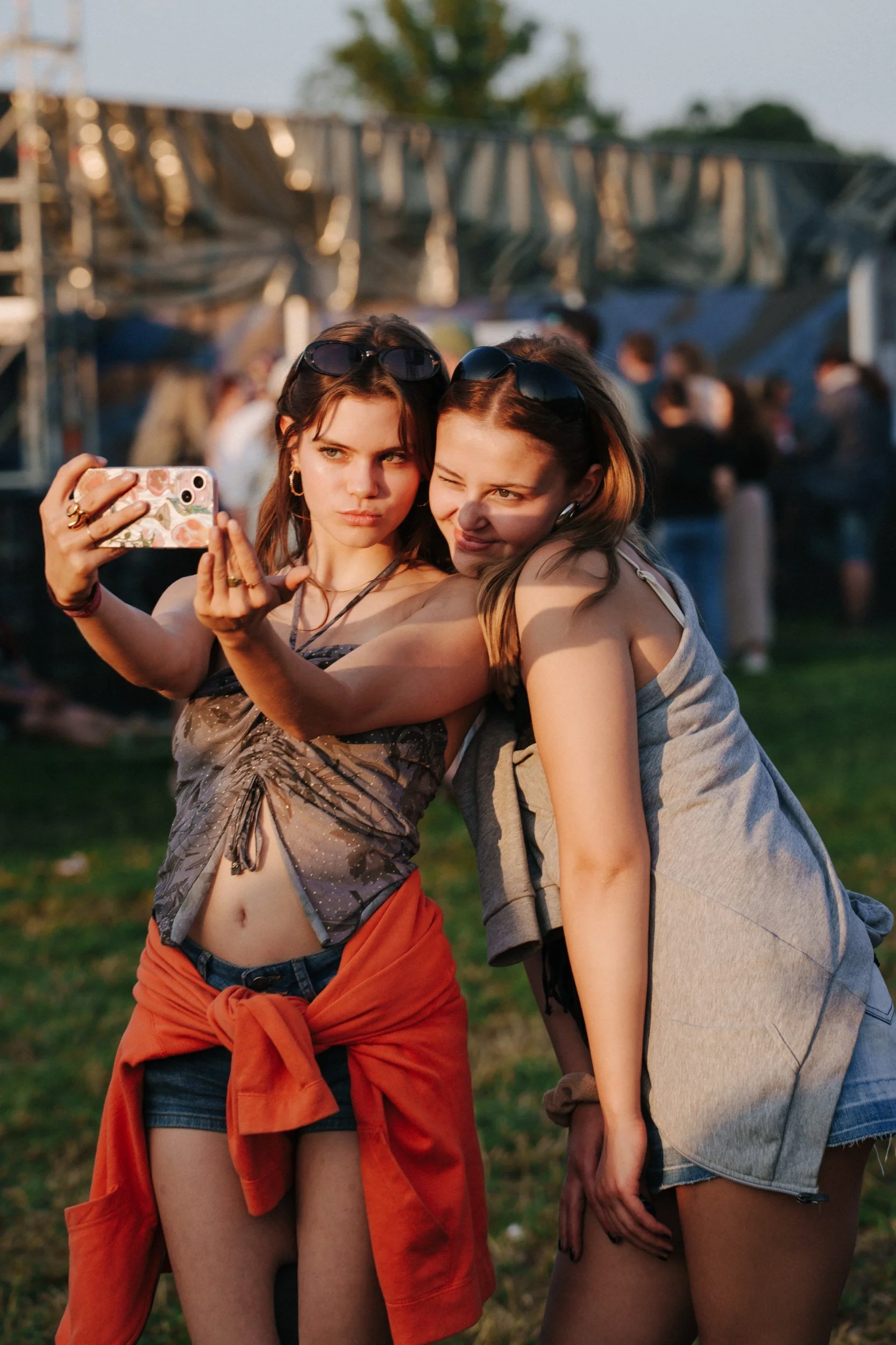 Zwei junge Frauen machen ein Selfie im Freien bei Sonnenuntergang, umgeben von anderen Menschen auf einem Festivalgelände.