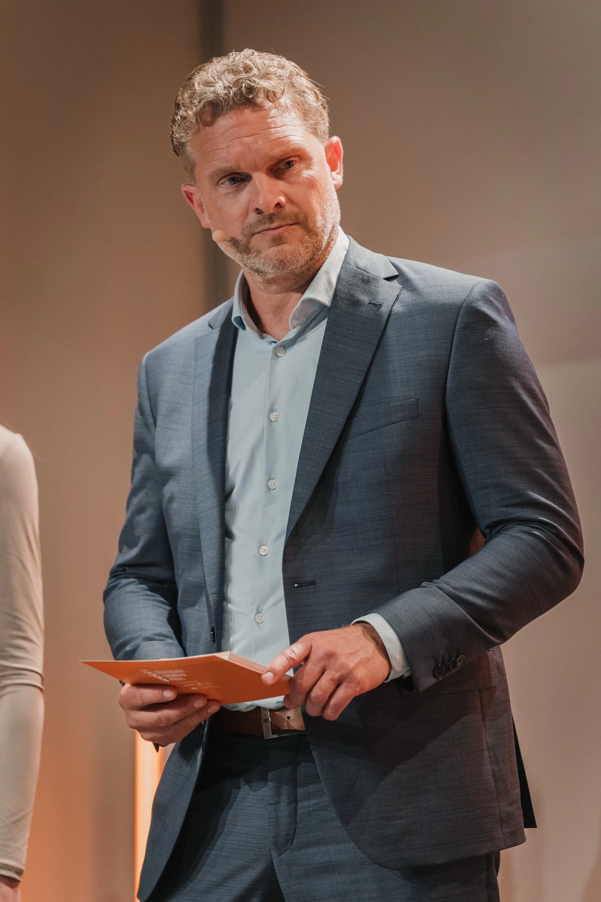 Ein Mann in grauem Anzug hält ein orangefarbenes Heft und schaut nach rechts.