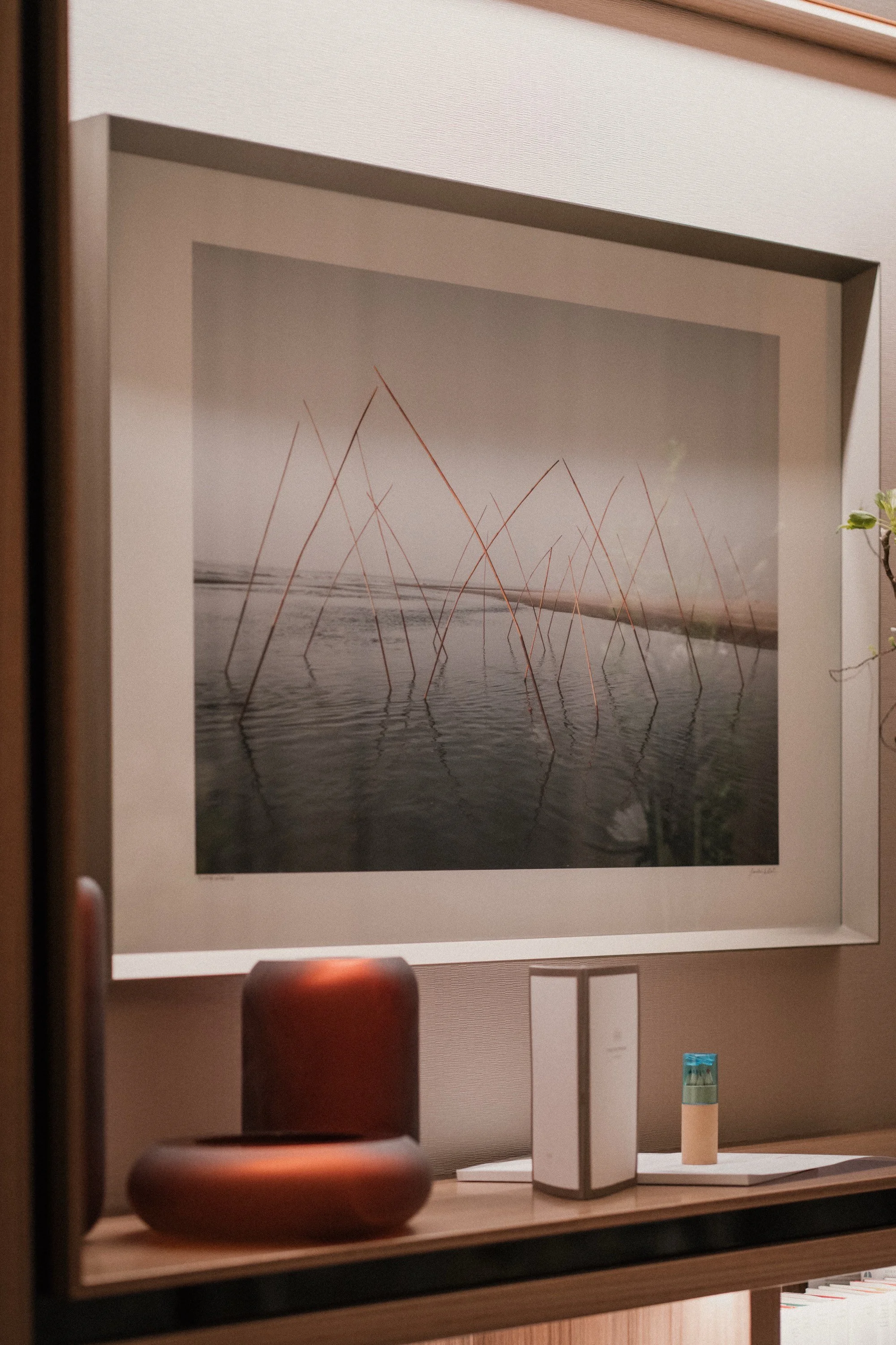 Ein gerahmtes Poster mit einer künstlerischen Fotografie von Wasser und dünnen, roten, gekrümmten Linien, die an Störche oder Vögel im Wasser erinnern, hängt an einer Wand in einem Raum. Unten auf der Ablage befinden sich ein brauner Keramikvase, ein