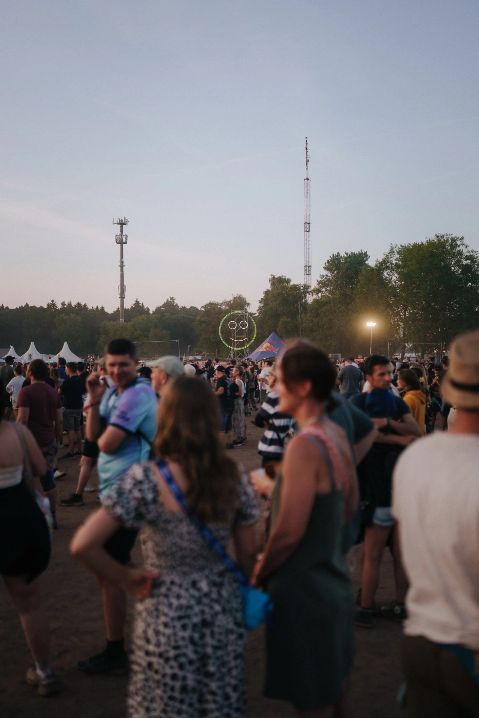 Menschenmenge auf einem Festival bei Sonnenuntergang mit Zufahrtszelte und einem lachenden Smiley am Himmel