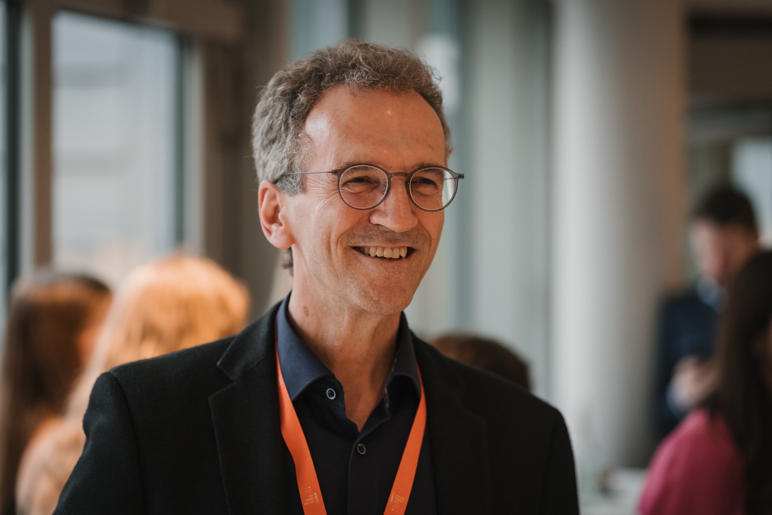 Ein lächelnder Mann mit Brille und dunklem Anzug bei einem Event in einem modernen Gebäude.