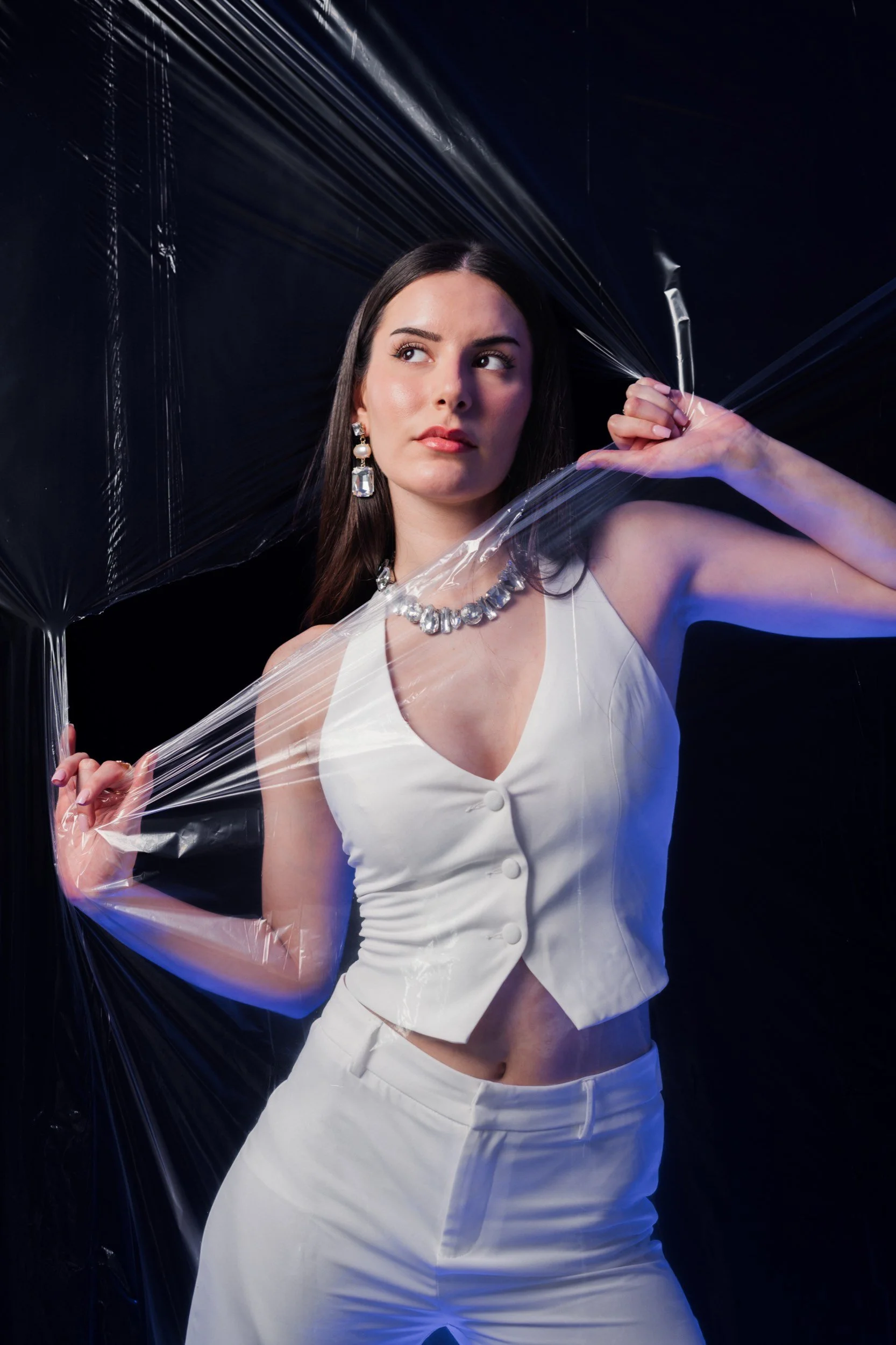 Junge Frau in weißem Outfit mit Schmuck posiert hinter transparentem Plastikfilm, dunkler Hintergrund.