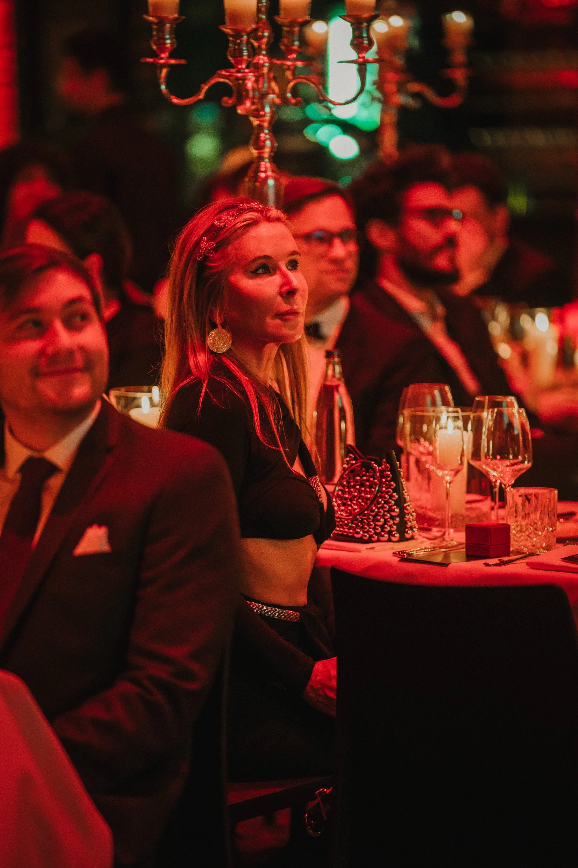 Menschen sitzen bei einer formellen Veranstaltung im abgedunkelten Raum mit roter Beleuchtung, bei Tischen mit Kerzen und dekorativen Gegenständen.