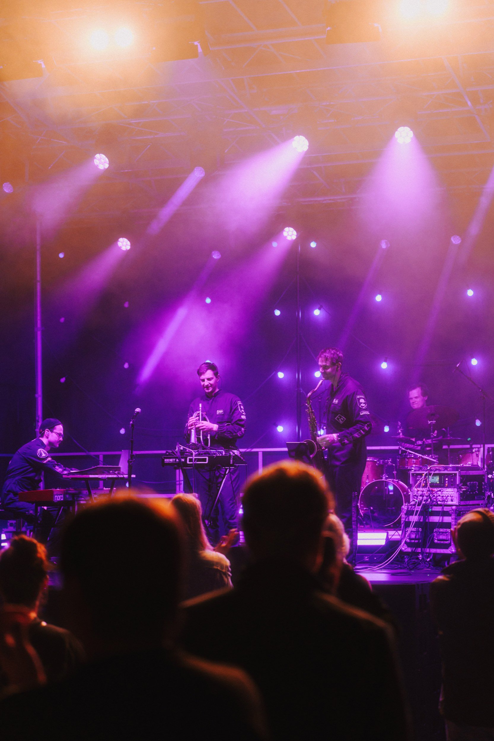 Eine Band spielt auf einer Bühne bei einem Konzert, beleuchtet mit pinken und violetten Lichtern, im Publikum sind Menschen zu sehen, die die Musik genießen.
