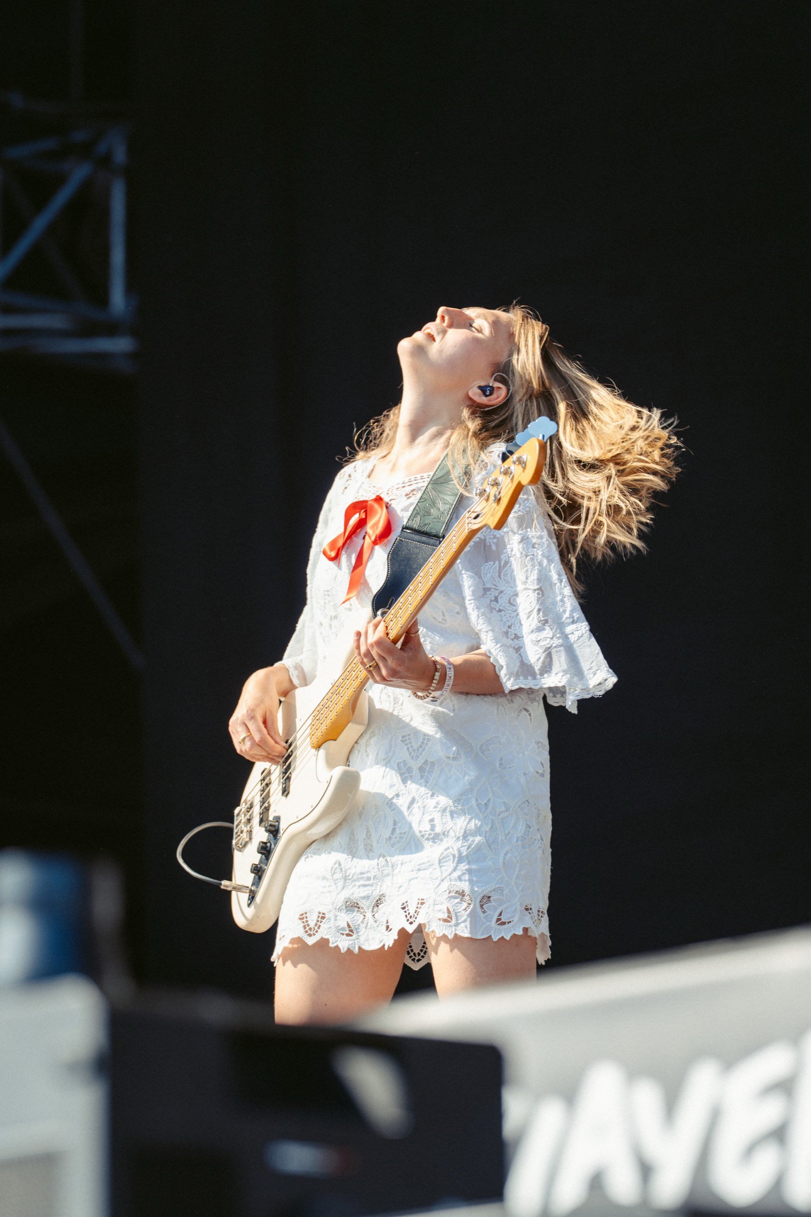 Junge Frau mit E-Gitarre beim Auftritt, trägt weißes Spitzenkleid und hat Locken, im Hintergrund dunkel.