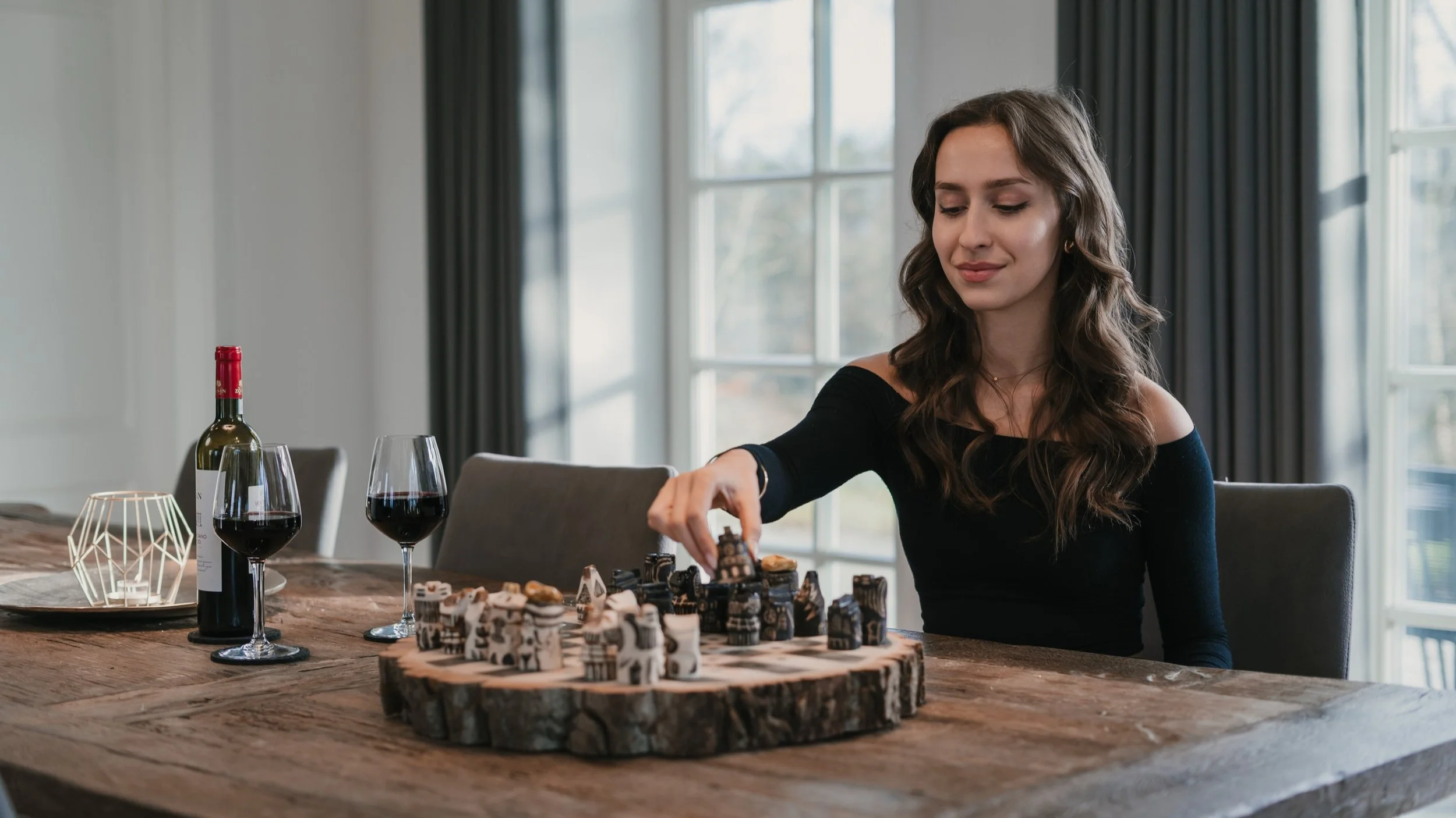 Junge Frau spielt mit Schachfiguren auf einem Holztablett, mit zwei Gläsern Rotwein und einer Weinflasche auf dem Tisch, im Hintergrund große Fenster.