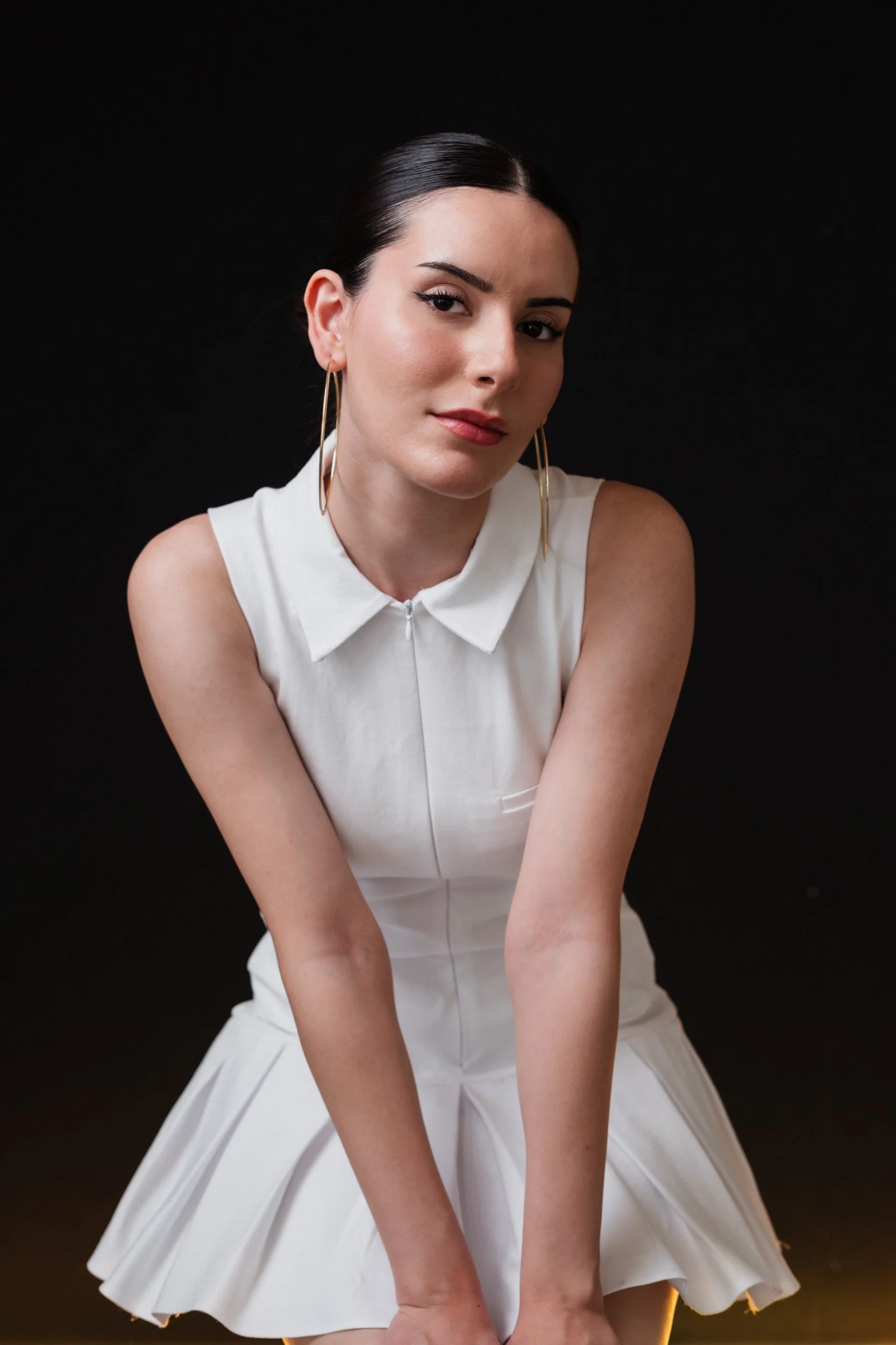 Junge Frau mit dunklen Haaren, weißen Kleid, großen Ohrringen gegen schwarzen Hintergrund.