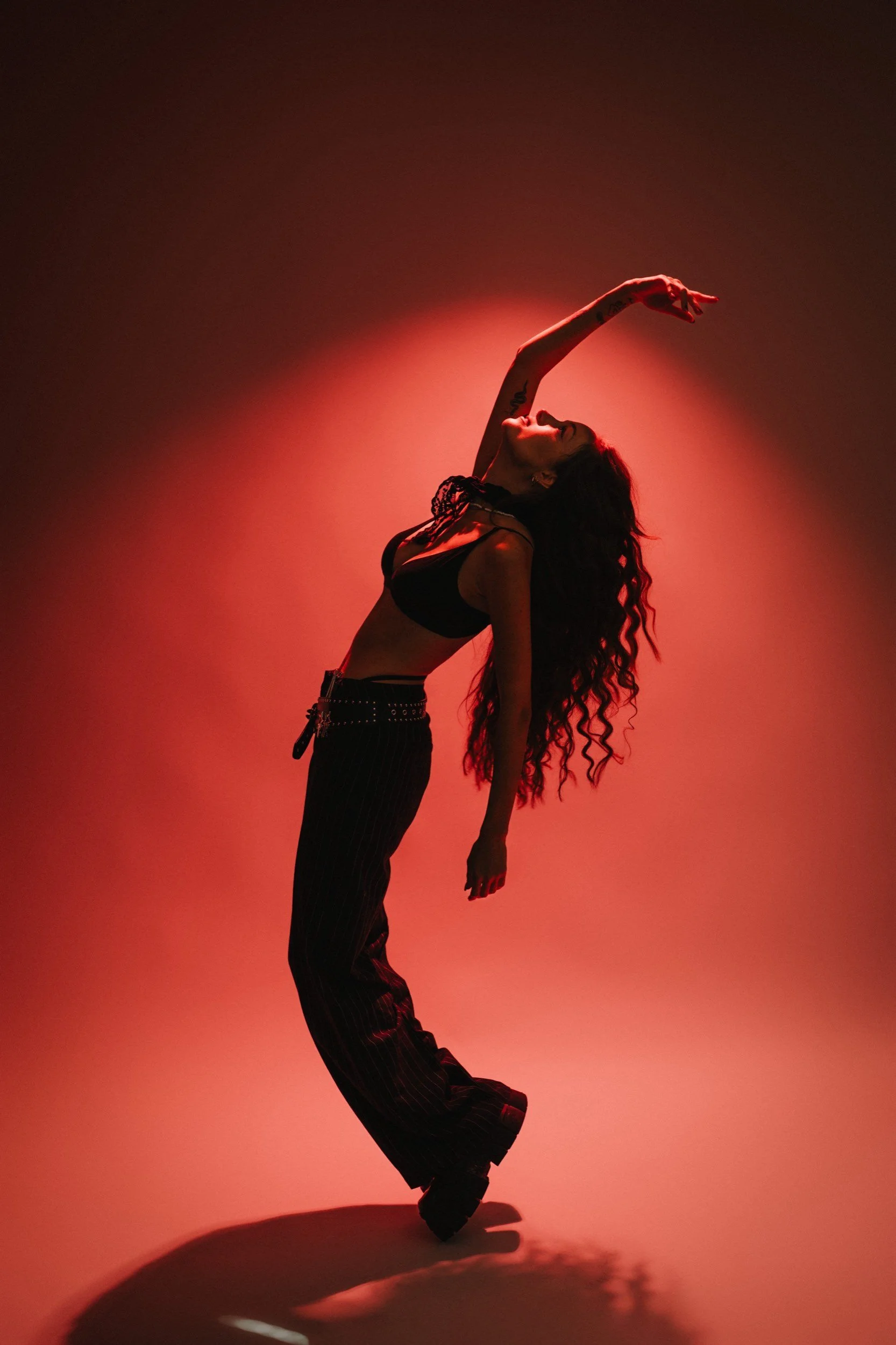 Eine Frau tanzt in einem schwarzen Outfit vor einem rot erleuchteten Hintergrund, mit einer dynamischen Pose.
