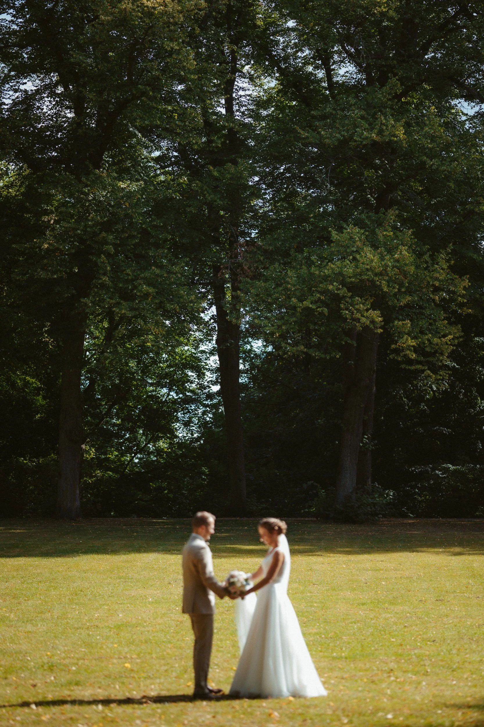 Ein Brautpaar steht im Freien auf einer Wiese, hält sich an den Händen und tauscht einen Blumenstrauß in einem Park mit großen Bäumen im Hintergrund.