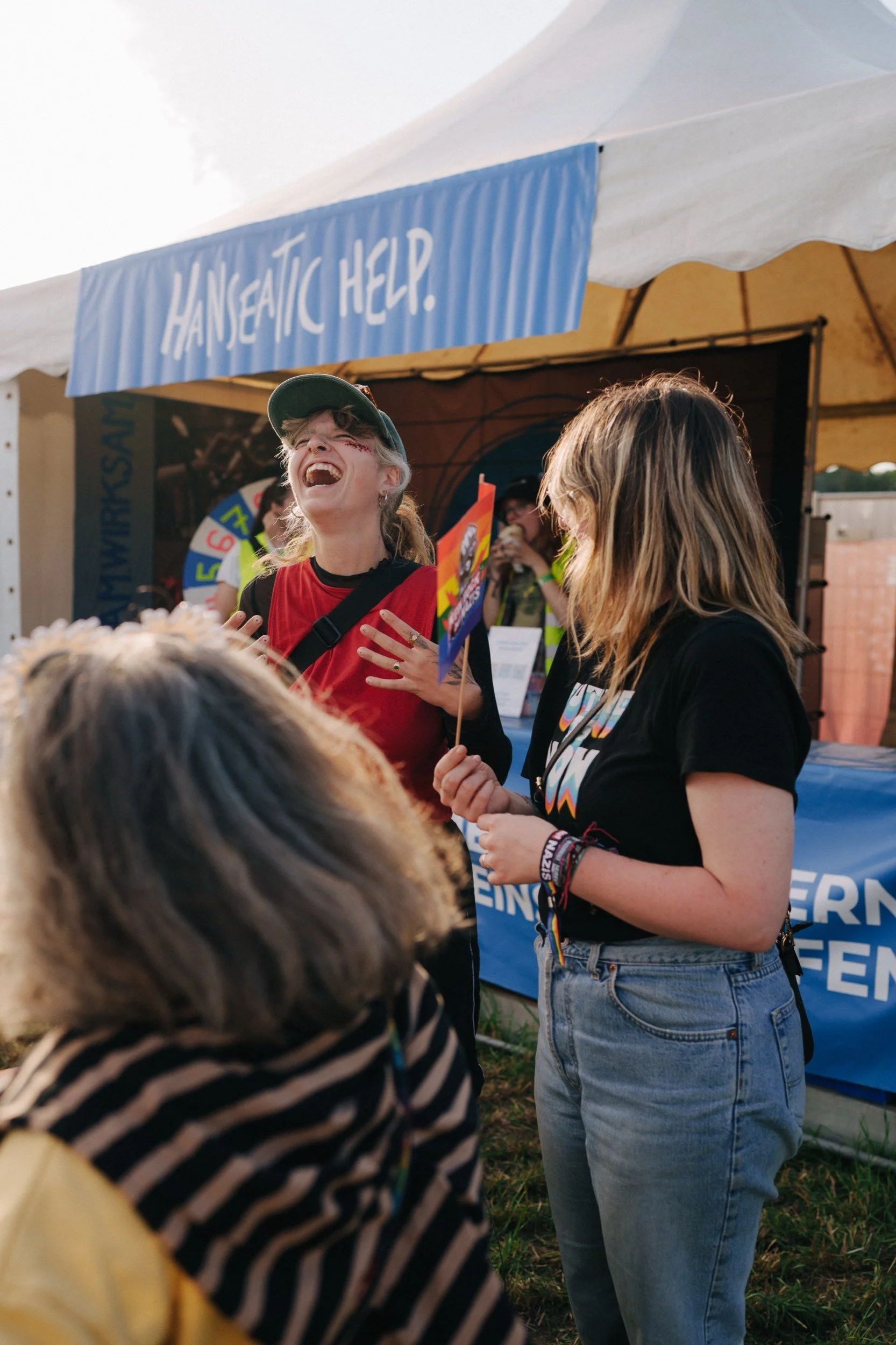 Junge Frau lacht herzlich bei einem Außenevent, während eine andere junge Frau mit Flagge neben ihr steht.