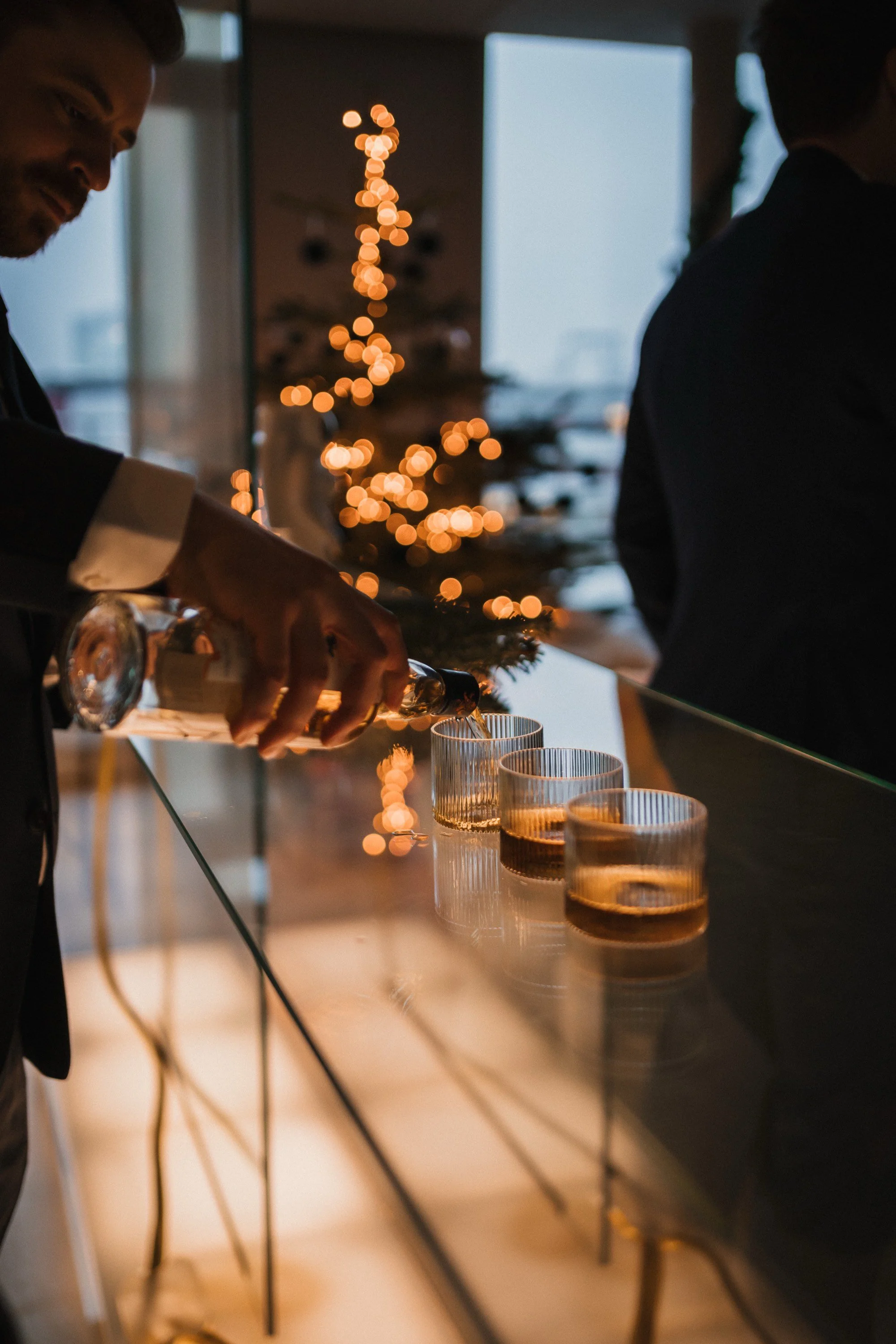 Ein Mann in einem Anzug gießt Getränke in Gläser an einer Bar mit einem Christmas Tree im Hintergrund.