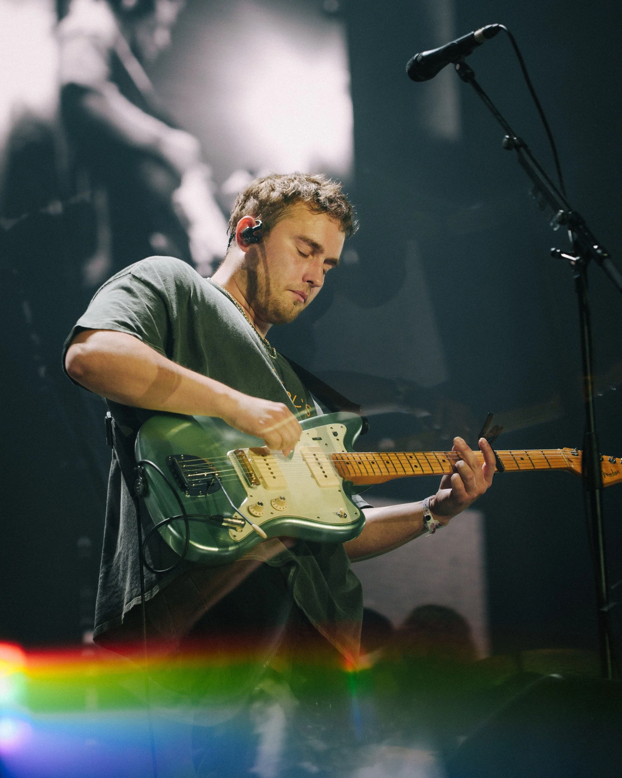 Junger Mann spielt Gitarre auf der Bühne bei einem Konzert, mit Hörmuscheln, dunklem Hintergrund und Mikrofon