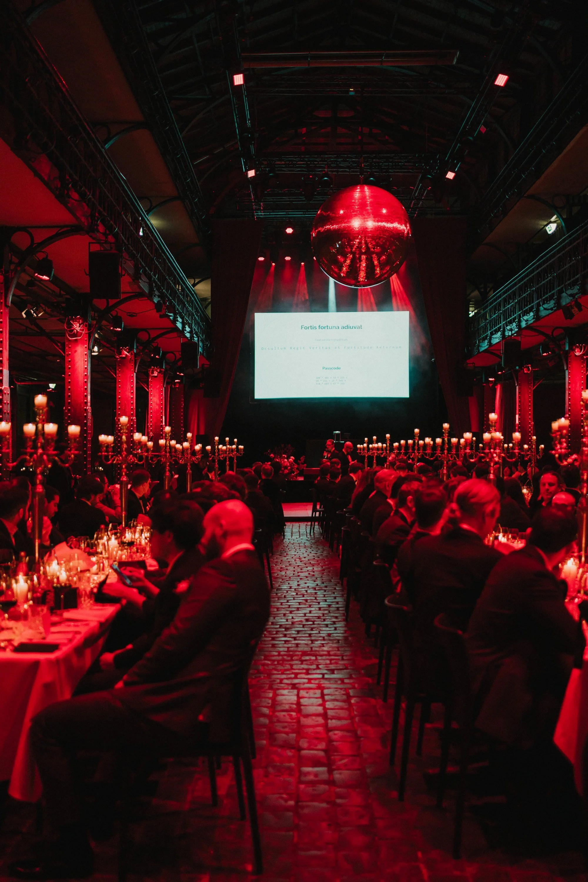 Ein eleganter Veranstaltungsraum mit roten Beleuchtungen, langen Tischen, Candle-Sticks und einem großen Bildschirm auf der Bühne. Es sitzen viele Gäste in dunkler Kleidung und der Raum hat eine historische, industrielle Architektur.