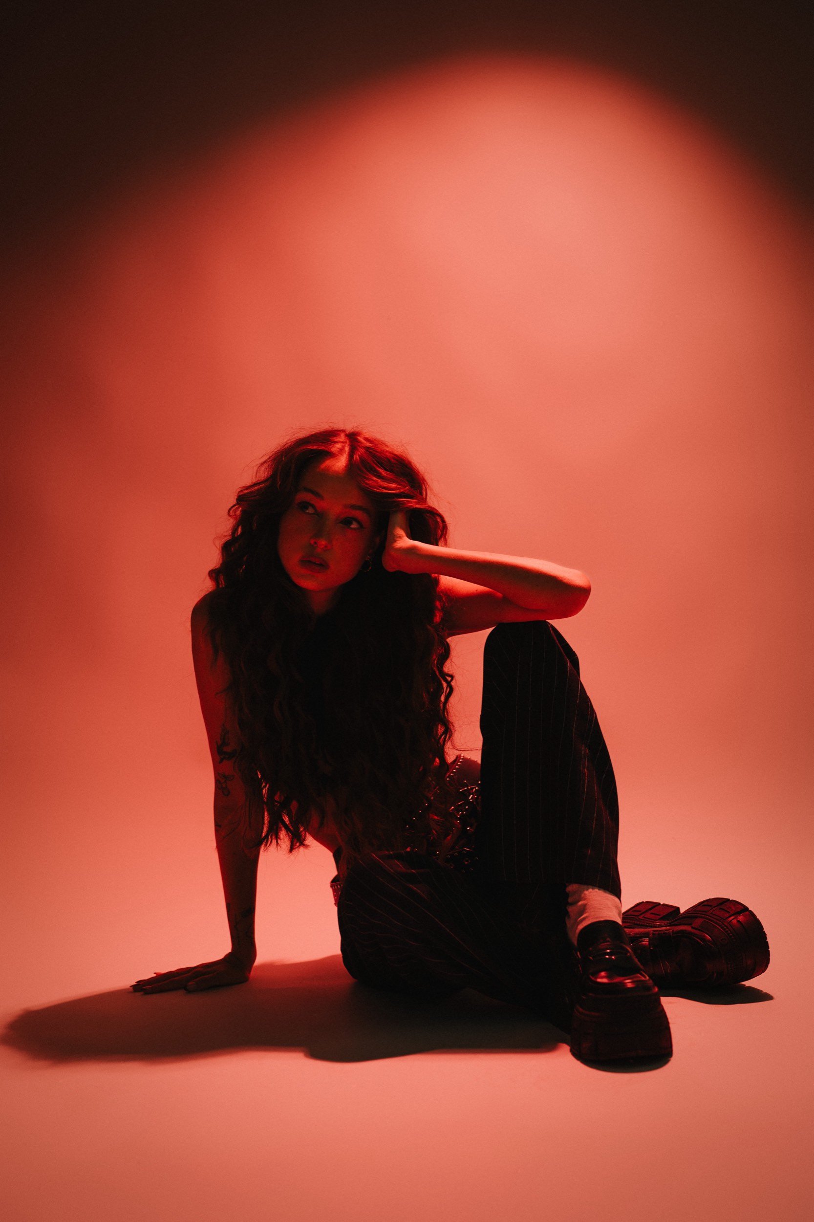 Junge Frau mit langen, lockigen Haaren sitzt auf dem Boden vor rotem Hintergrund, die Hand an den Kopf gelegt, trägt schwarze Schuhe und gestreifte Hose.