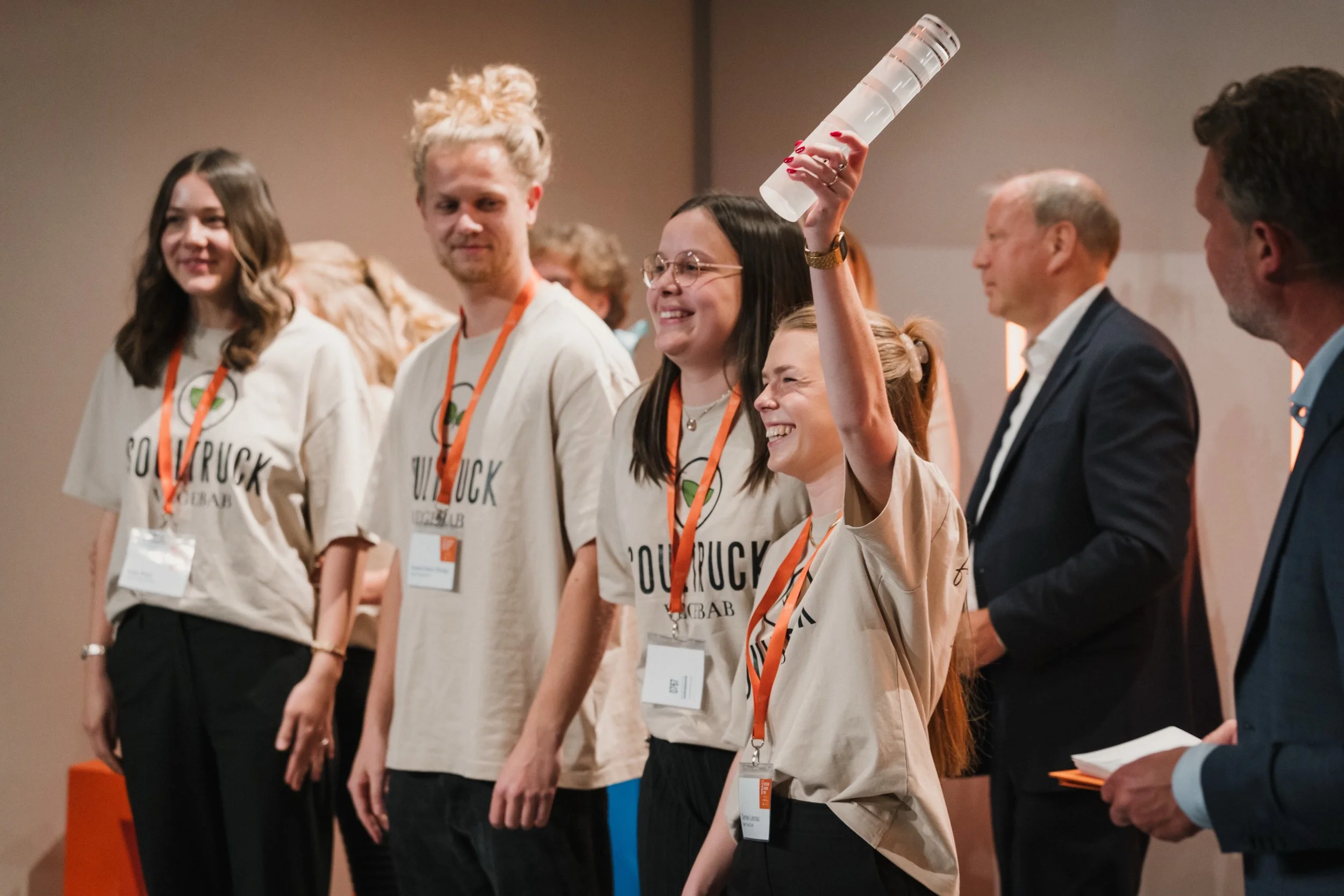 Gruppe von Menschen auf einer Bühne, vier junge Frauen tragen beige T-Shirts mit Aufdruck, eine Frau hebt ein Glas in die Luft und lächelt, zwei Männer in Anzügen stehen daneben.