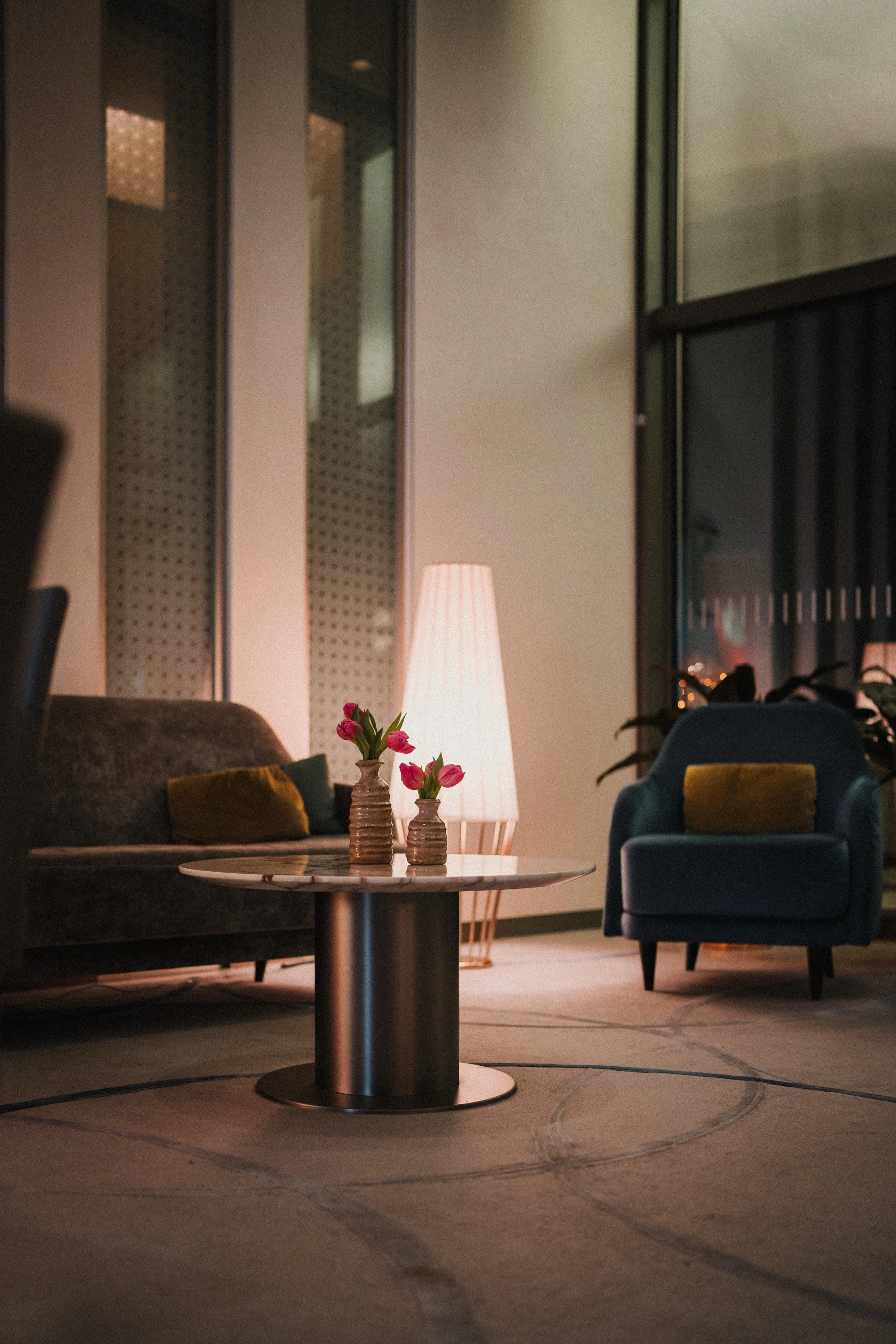 Gemütliches Wohnzimmer mit einem runden Couchtisch, zwei Vasen mit pinken Blumen, einem Stehlampen, einem grauen Sofa mit Kissen und einem blauen Sessel, große Fenster im Hintergrund.