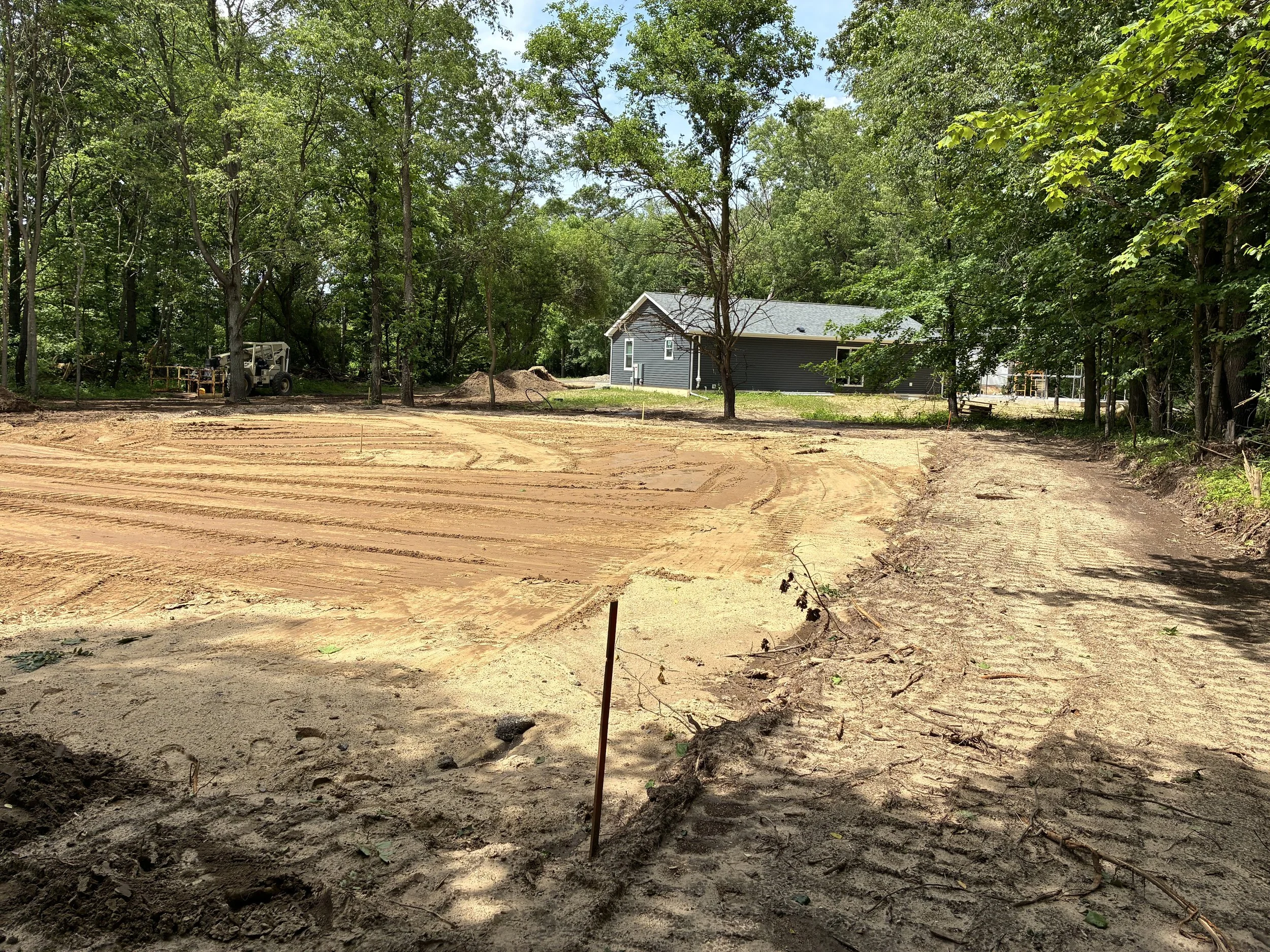 A construction site with cleared dirt, trees surrounding, and a house being built in the background, with construction equipment present.