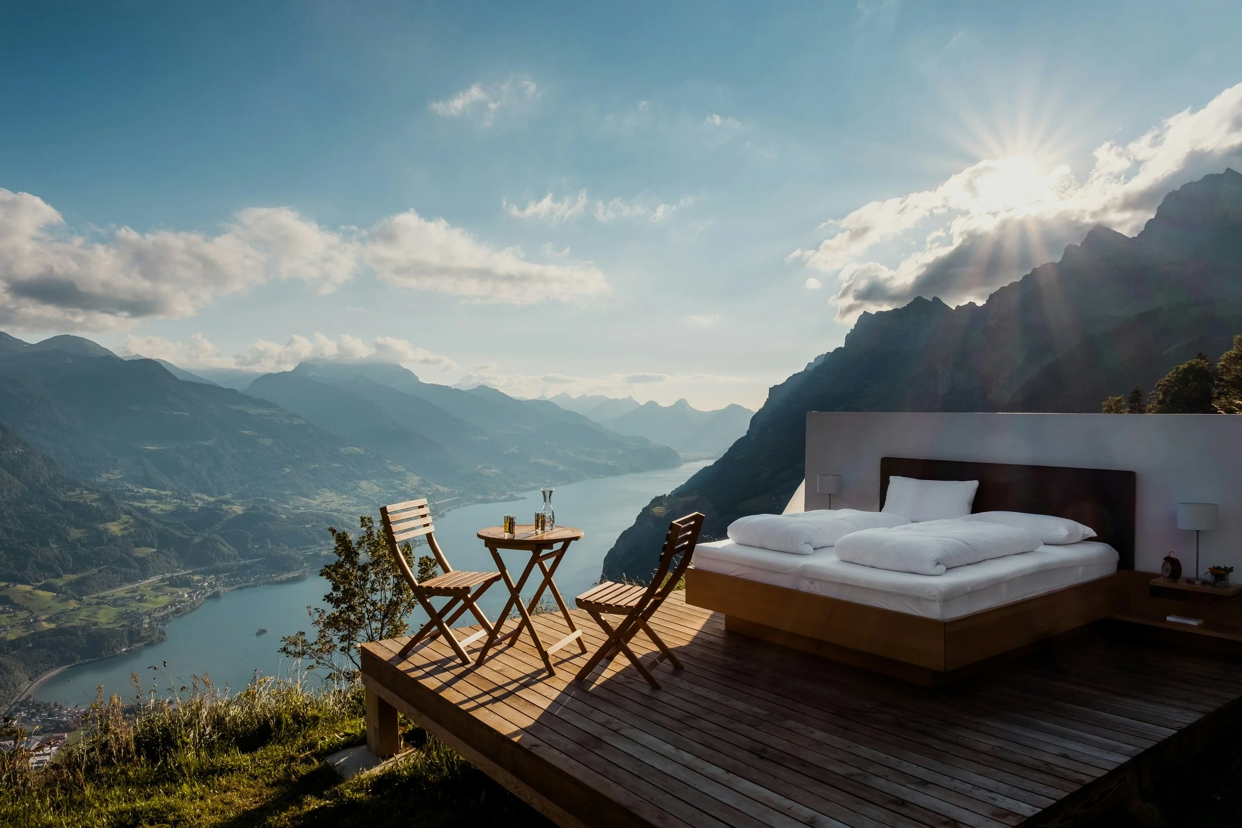 An outdoor wooden deck with a bed, two table lamps, and a small round table with two chairs, overlooking a valley with a river surrounded by mountains and a partly cloudy sky with the sun shining.