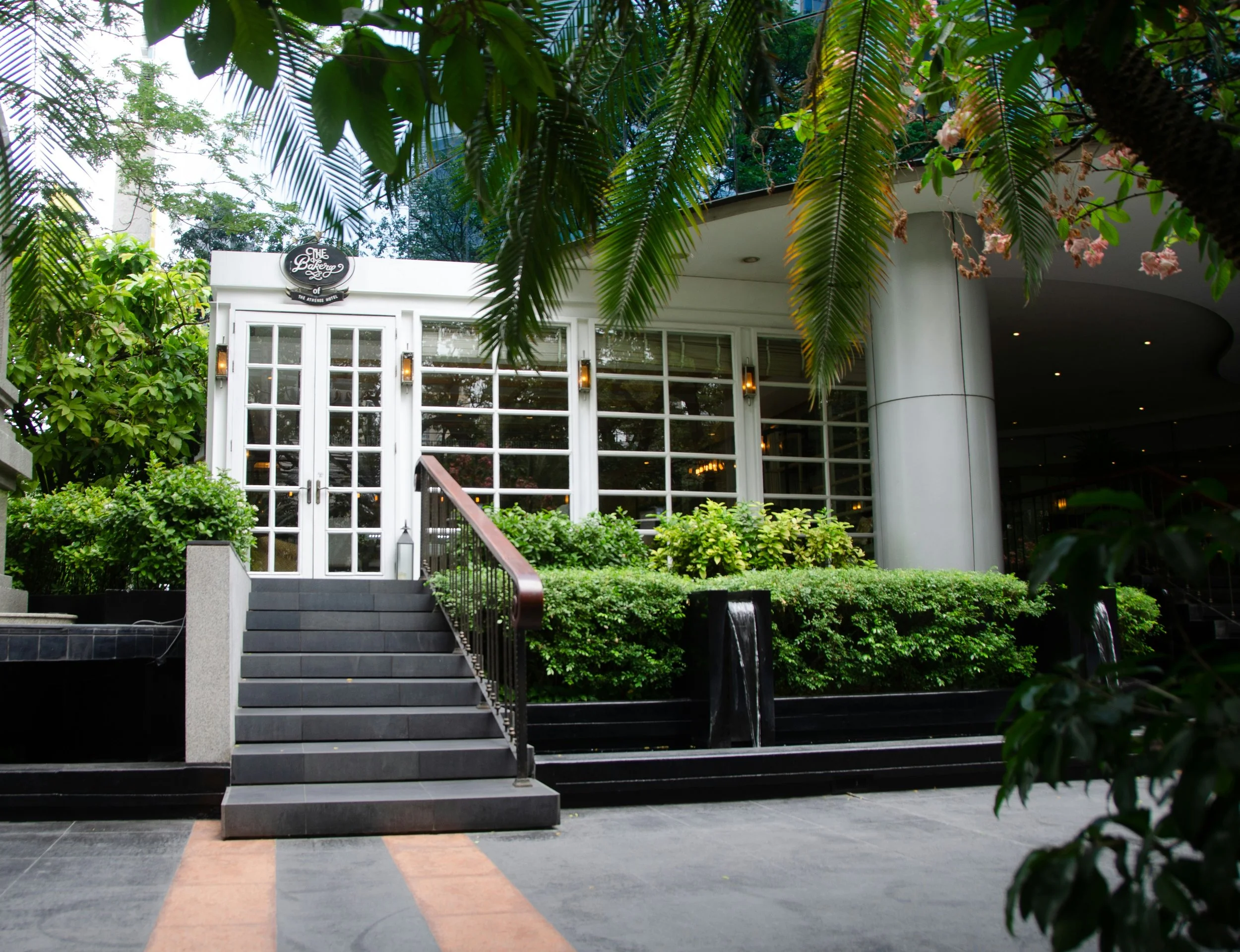 Entrance of a restaurant or cafe with white glass doors, surrounded by green bushes and plants, with black stairs leading up to the door, framed by trees and greenery.