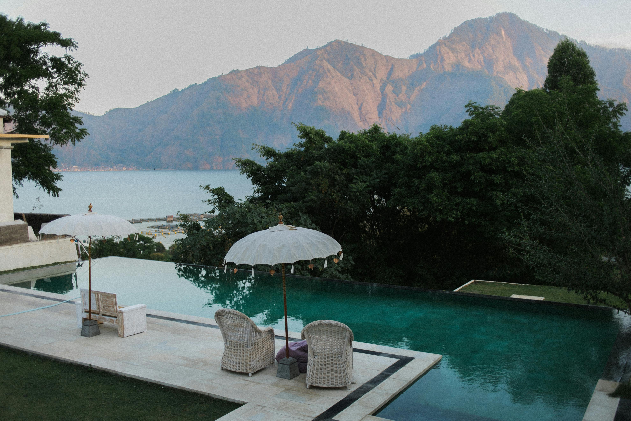 View of a luxurious infinity pool with two wicker chairs and umbrellas, overlooking a lake and mountains in the distance during dusk.