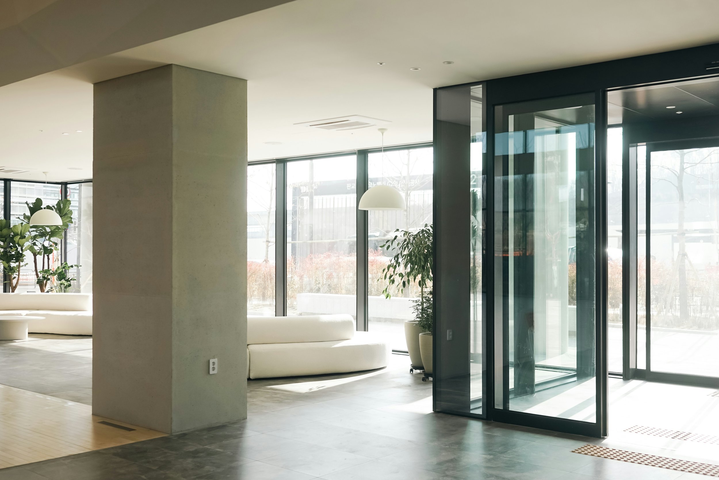 Modern lobby with large floor-to-ceiling glass windows, white curved sofas, potted plants, and a sliding glass door entrance.