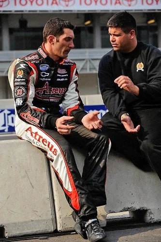 Two men, one dressed in a racing suit and the other in black clothing, sitting and talking at a racing track.