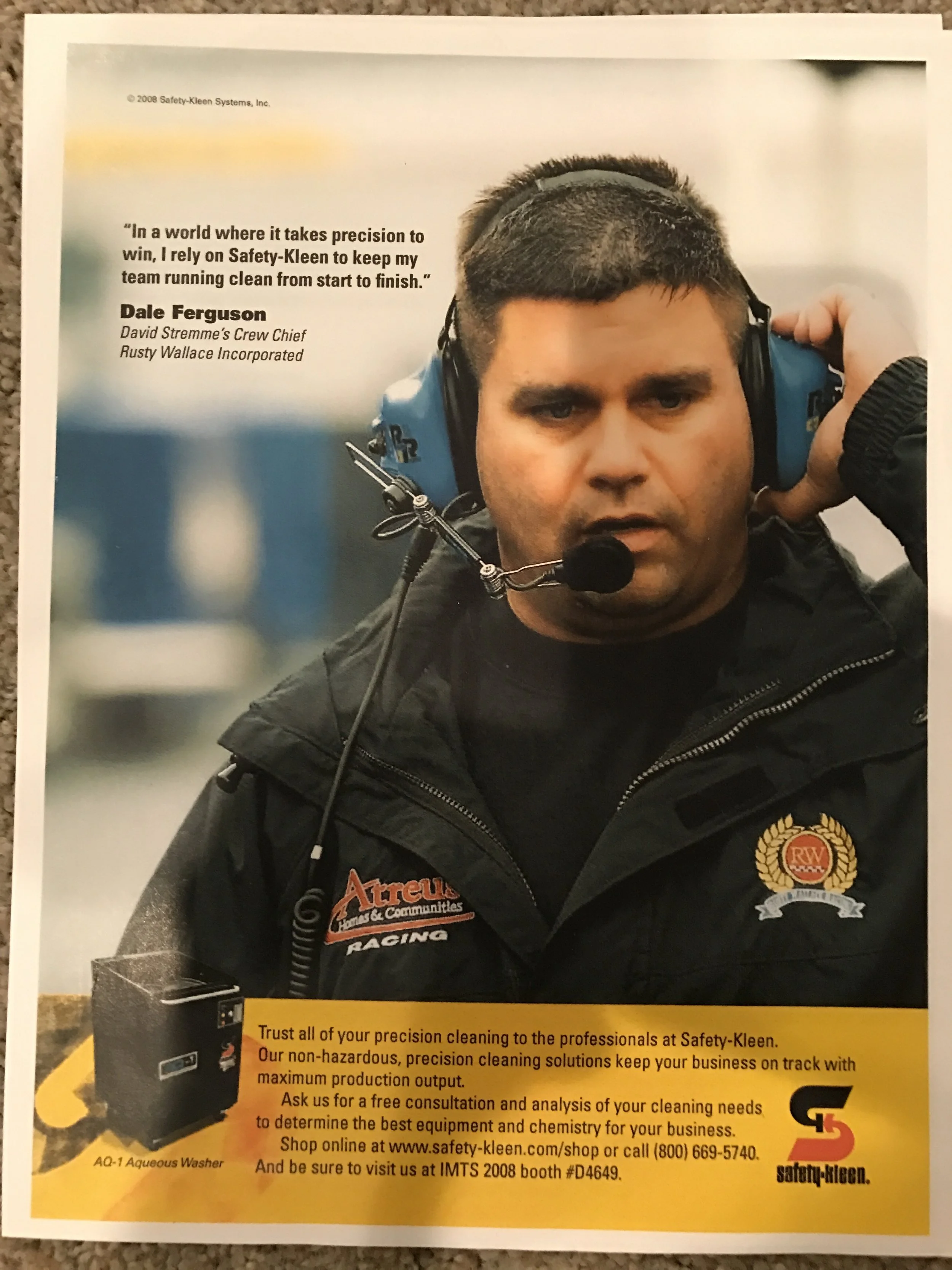A man in a black jacket with safety-based patches, wearing blue noise-canceling headphones with a microphone, is looking into the camera.