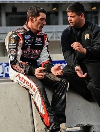 A race car driver in a black and red racing suit sitting on a concrete barrier, talking to a man in a black jacket who is gesturing with his hands. They are at a racetrack.