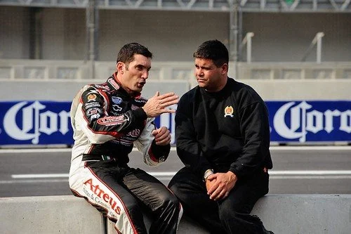 Two men engaged in a discussion at a racing track, one wearing a racing suit and the other in a black jacket, with a Corona advertisement in the background.