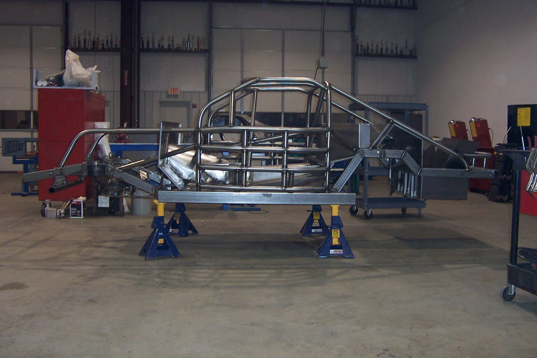 Metal framework of a race car chassis being assembled on blue stands in an industrial workshop.