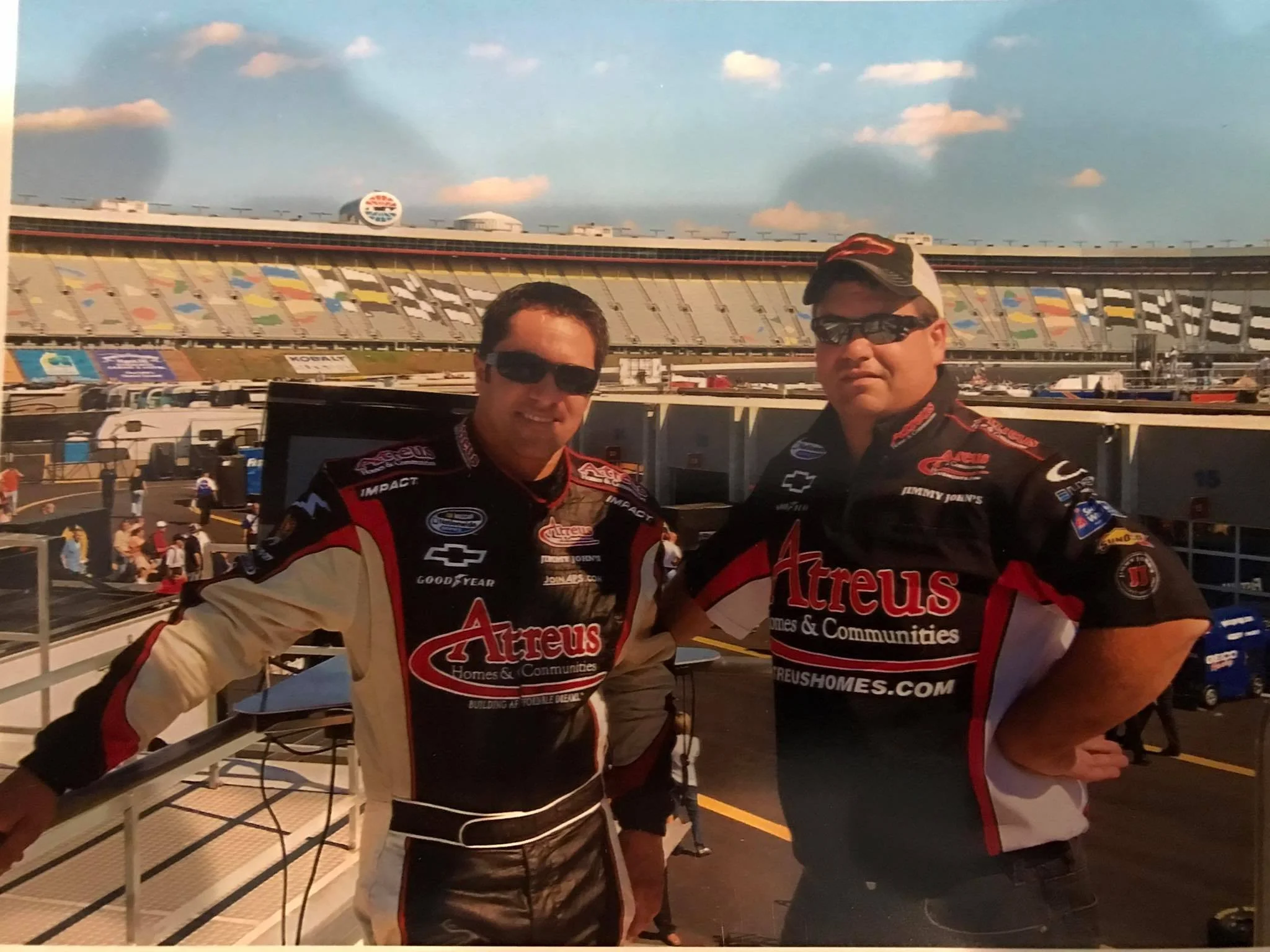 Two men in racing suits smiling at the camera at a race track, with empty grandstands and a partly cloudy sky in the background.