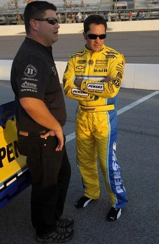 Two men standing on a race track, one in a black shirt and the other in a yellow racing suit with sponsor logos, both wearing sunglasses.
