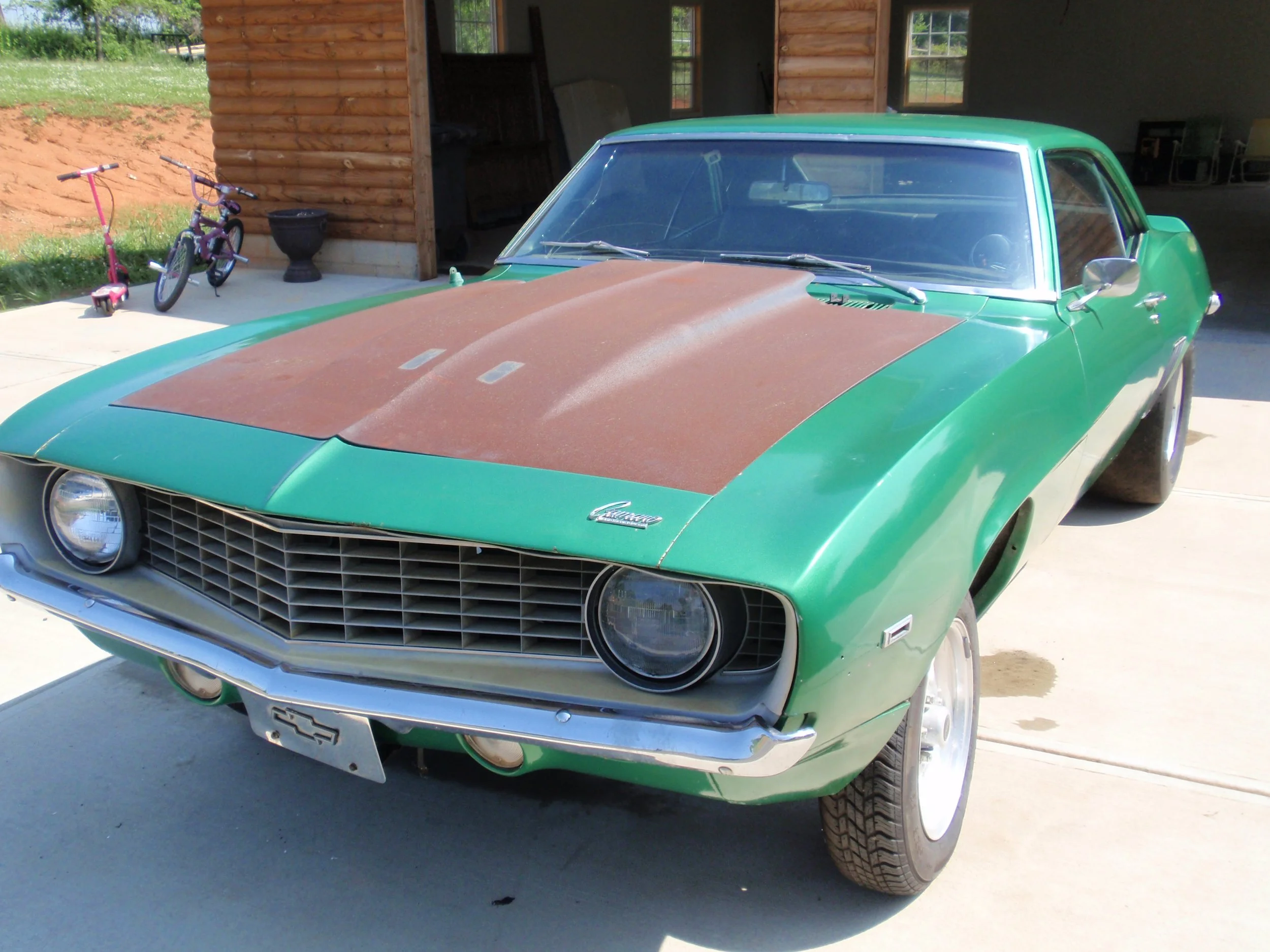 A vintage green Chevrolet Camaro car with a rust-colored hood parked in front of a wooden building, with a small pink and purple children's bicycle and a pink scooter nearby.