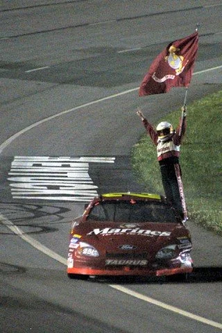 A person standing through the sunroof of a red race car, waving a flag on a racetrack.