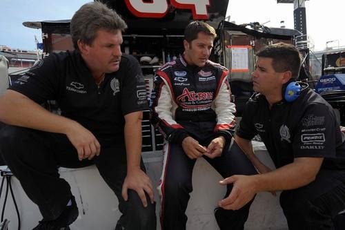 Three men sitting and talking in a racing garage area, with race car equipment visible in the background.