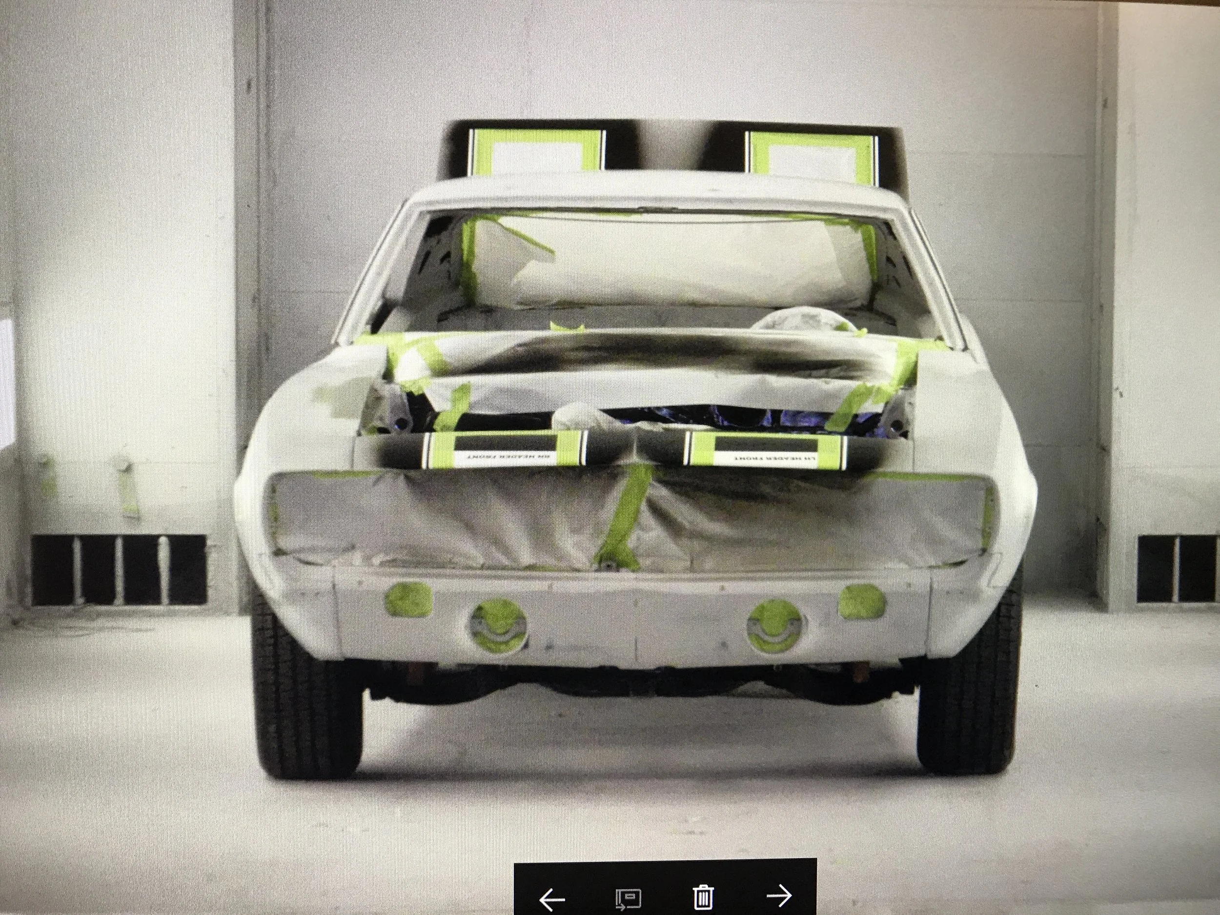 A model or toy of a white car with green tape on various parts, placed in a room with white walls and a vent on the left side.
