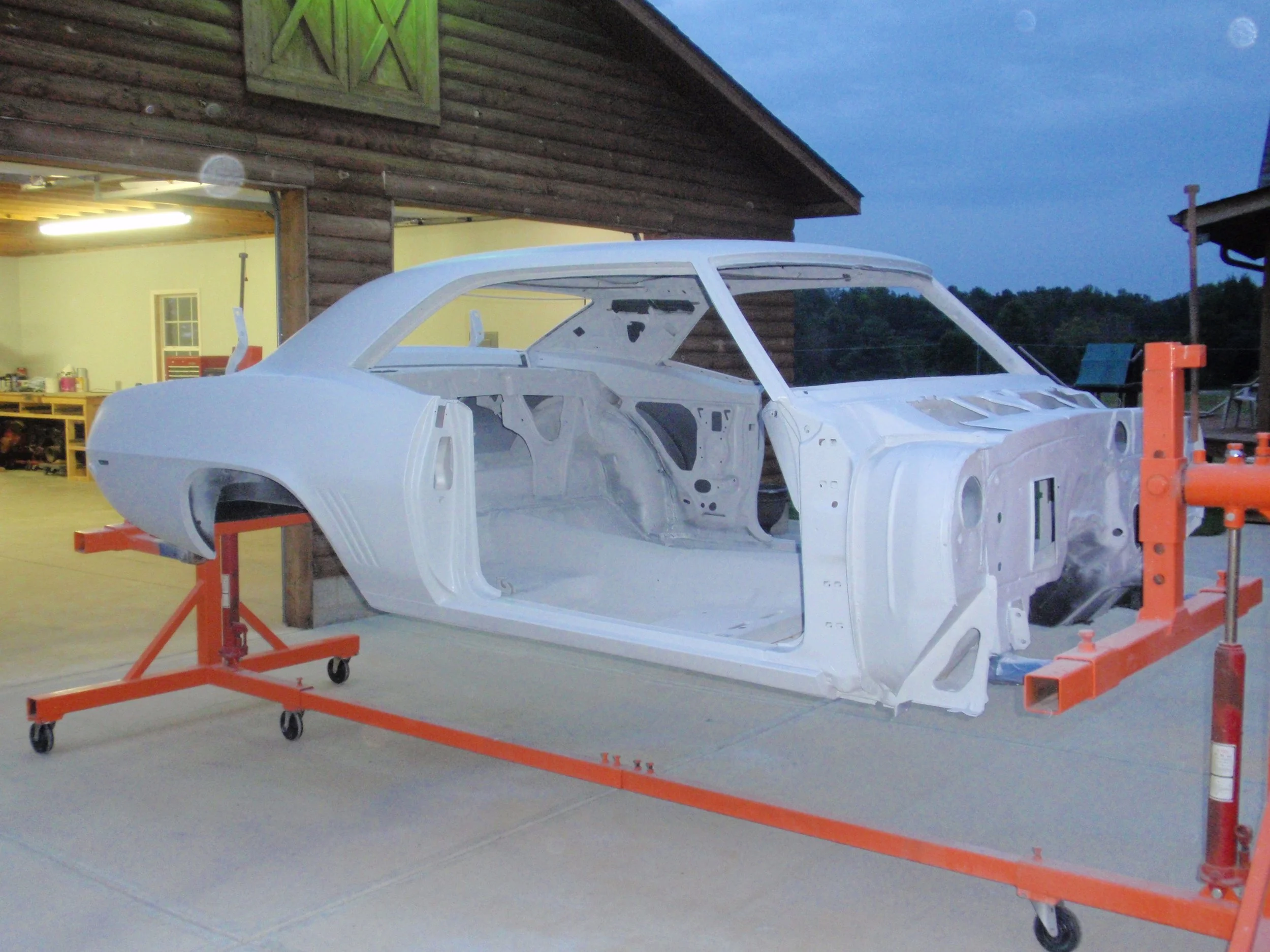 A white car body shell on an orange rolling cart inside a garage or workshop. The background shows a wooden building exterior with a window and an open garage door revealing tools and supplies. The scene appears to be during early evening or dusk.